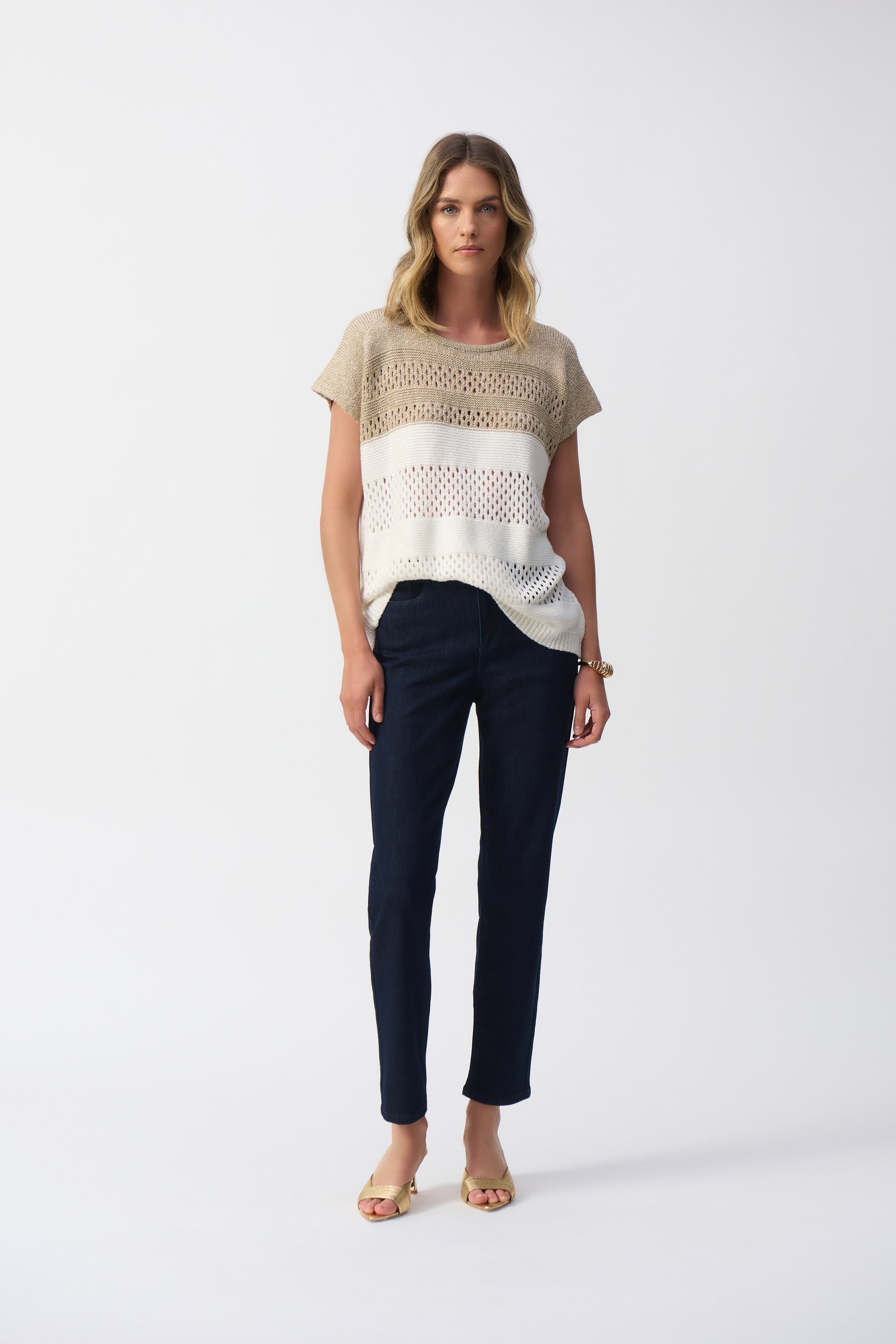 Woman wearing a beige and white colour block knit sweater with short dolman sleeves, paired with dark skinny jeans and gold open-toe heels, standing in a studio setting.