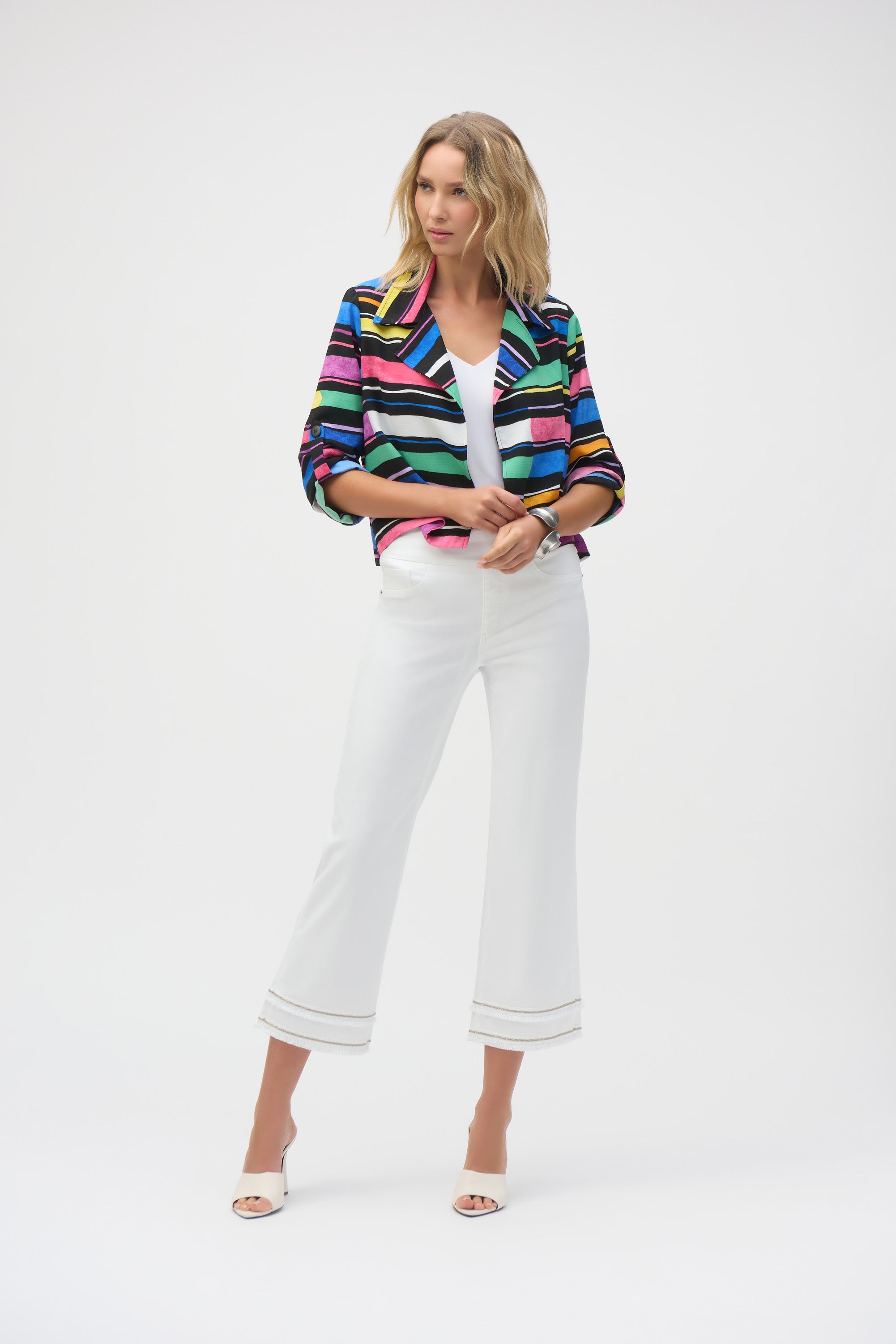 Woman posing in a studio wearing a colourful striped blazer and white cropped pants with fringe hem.