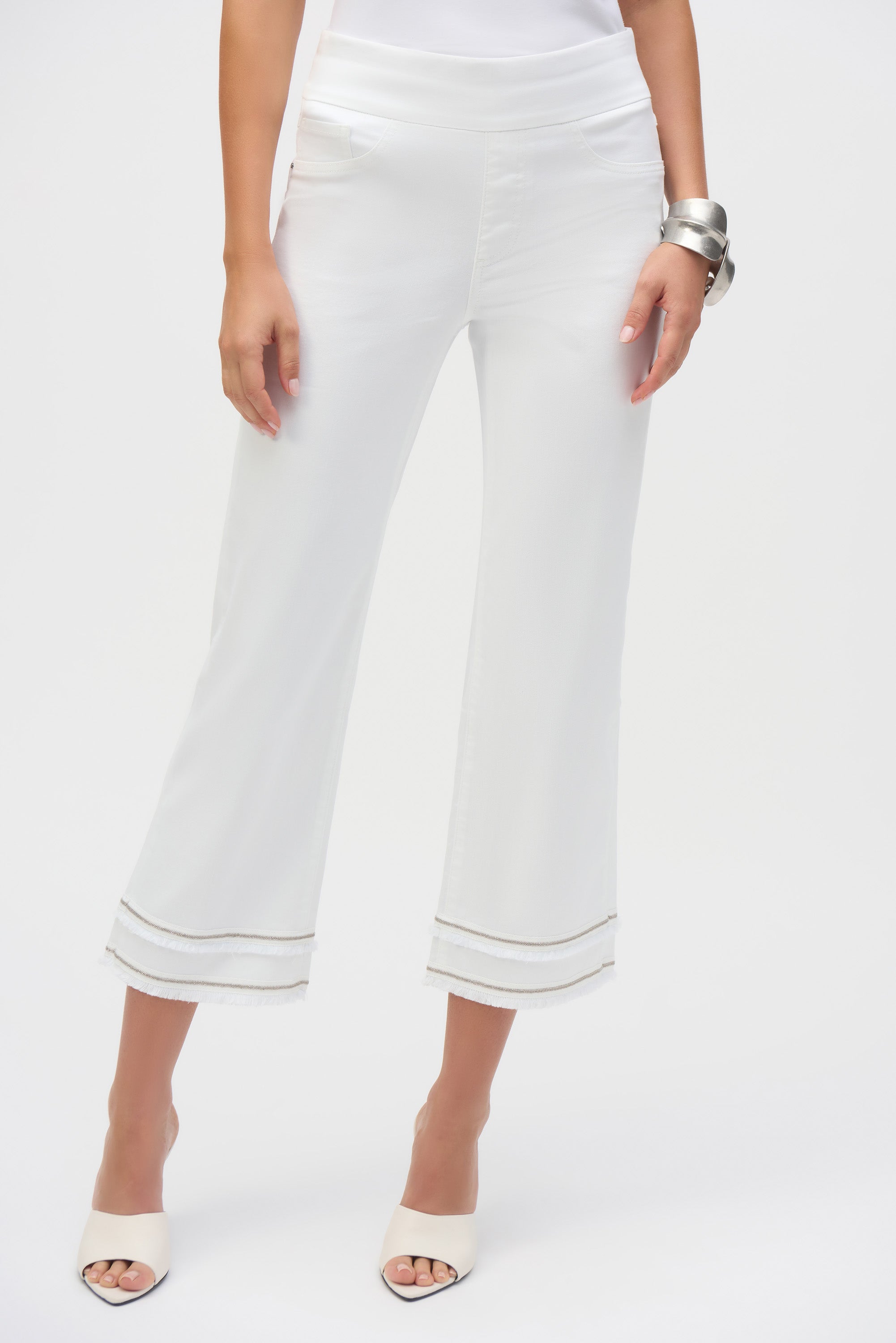 Front view of white cropped pants showing the clean waistband and fringe hem, styled with a white tank and silver bangles.