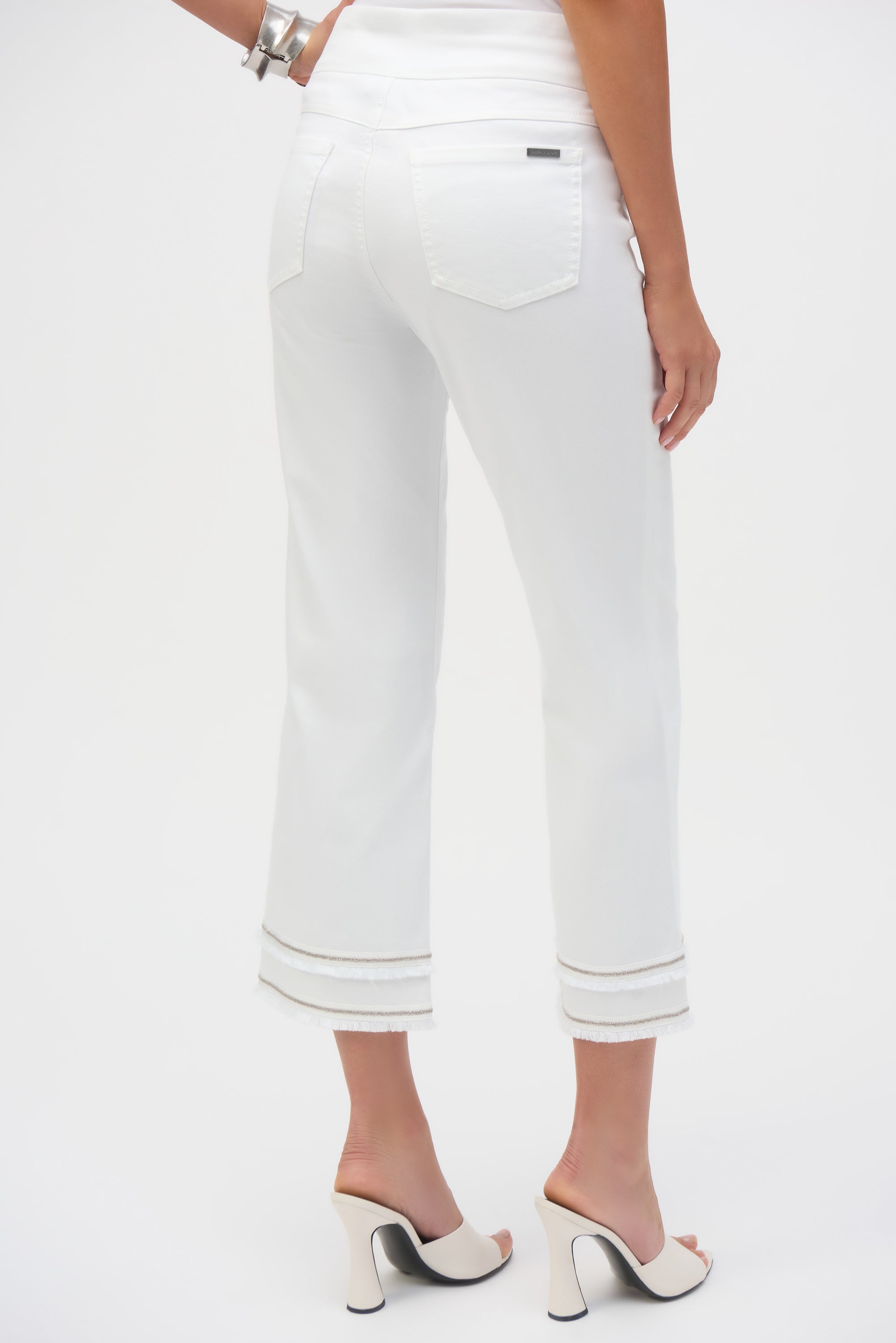 Back view of white cropped pants with patch pockets and fringe trim at the hem, worn with white heels.