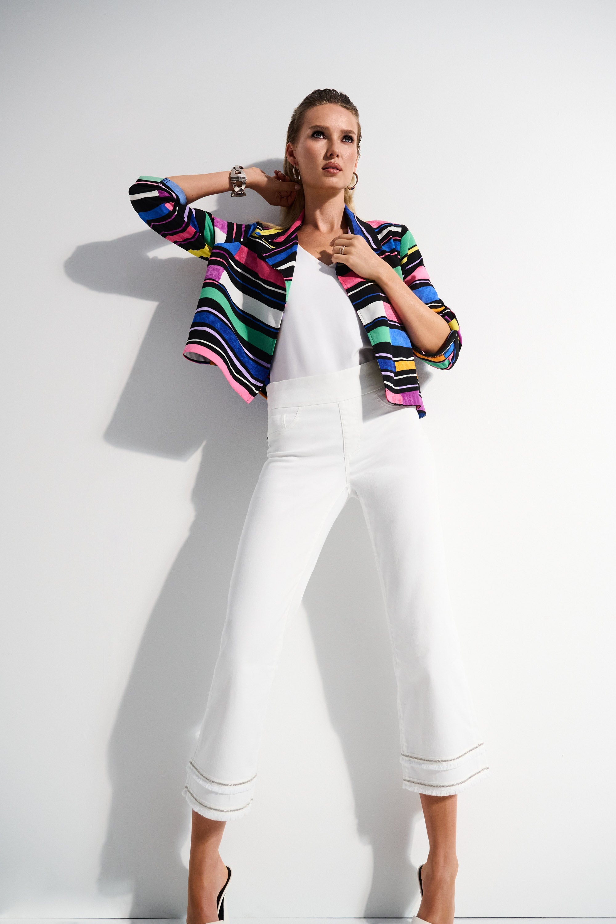 Woman wearing colourful blazer and white cropped pants styled with white heels, posing against a white backdrop.