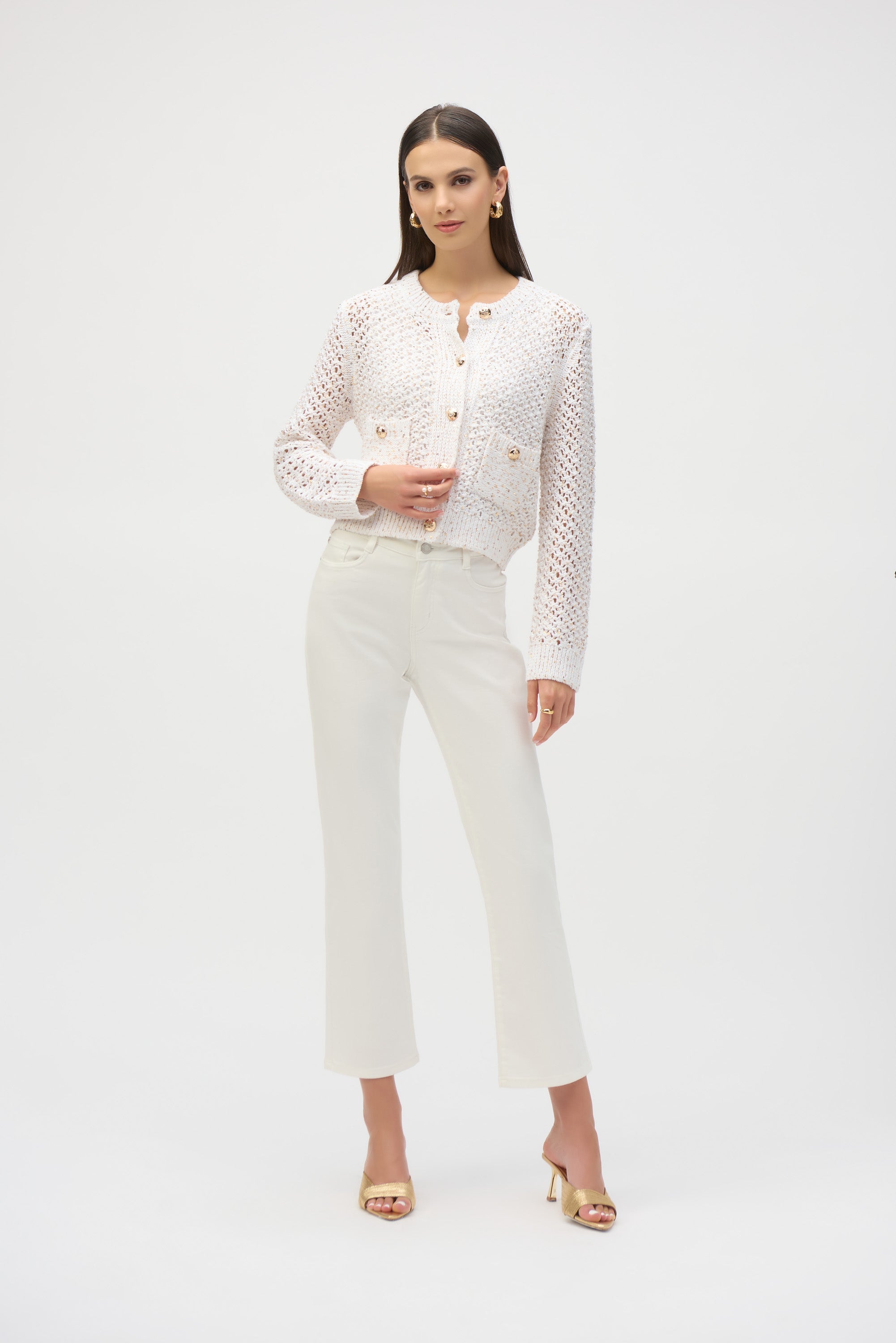 Full-length view of woman styled in cream chenille open stitch cardigan with white cropped pants and gold heels.