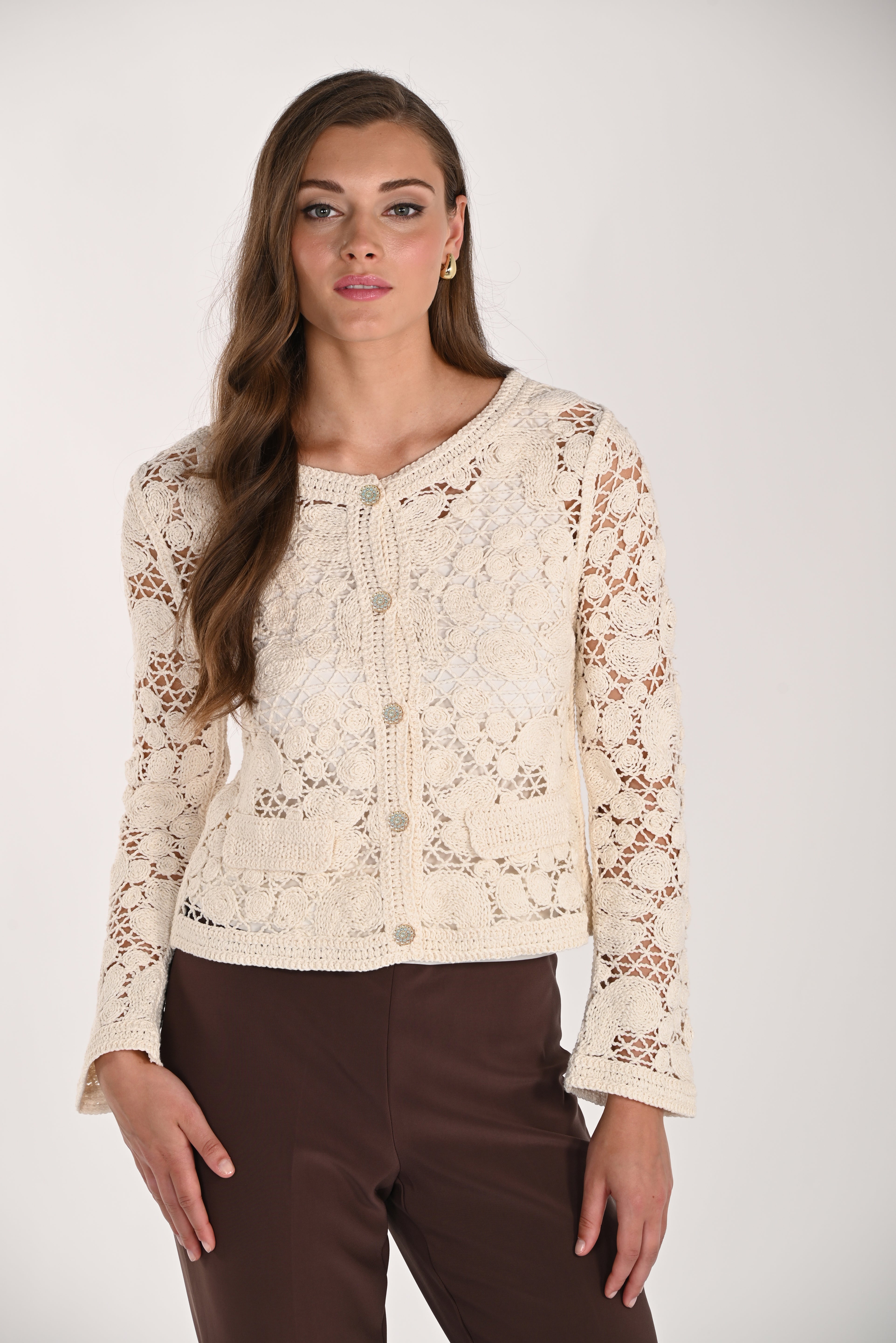 Model wearing a cream crochet cardigan featuring floral crochet detailing, button-front closure, long sleeves, and front patch pockets paired with brown trousers.
