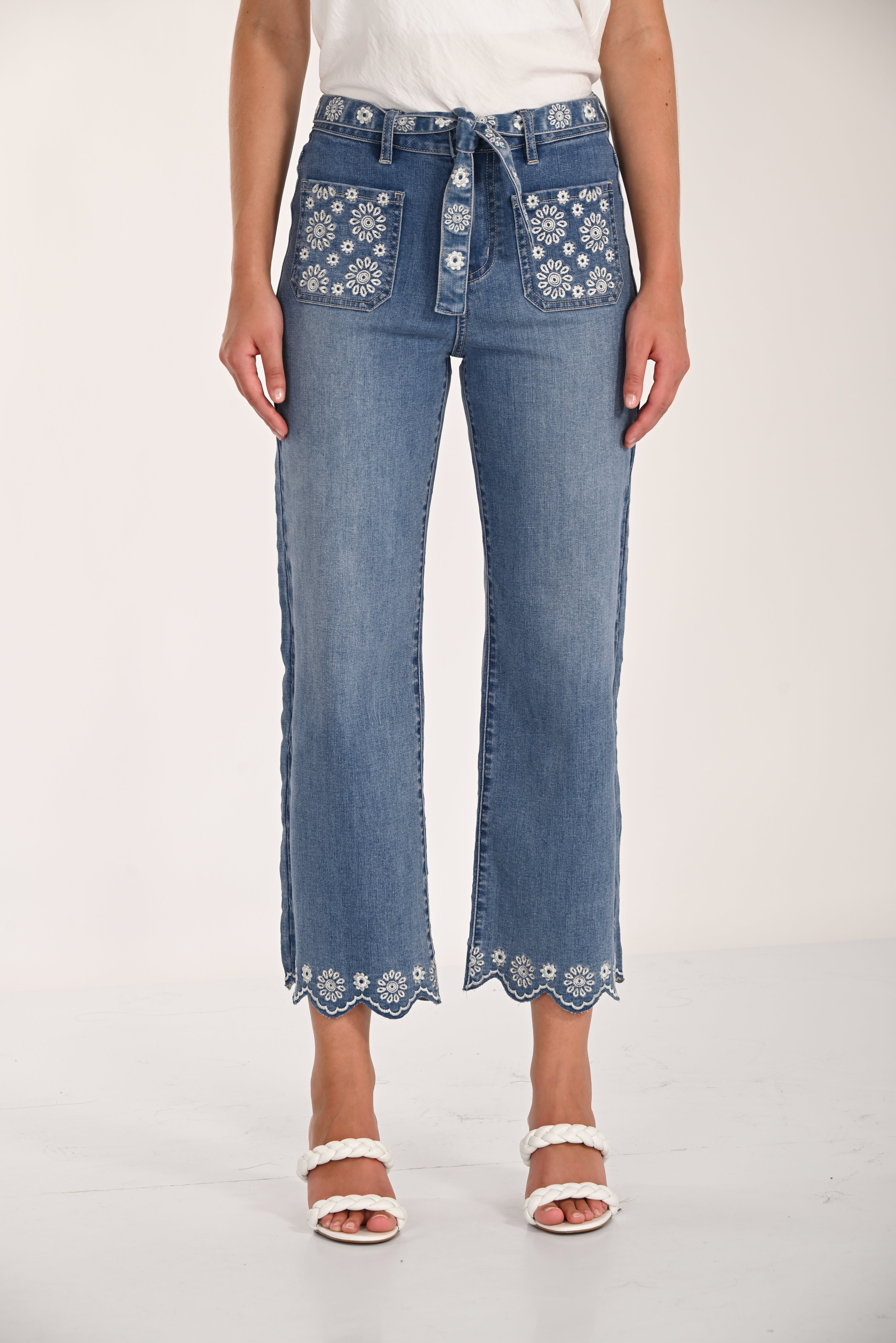 Woman wearing medium-wash embroidered cropped wide-leg denim pants with a tie waist, floral embroidery on the front pockets and waistband, and scalloped embroidered hems, styled with white braided heeled sandals.