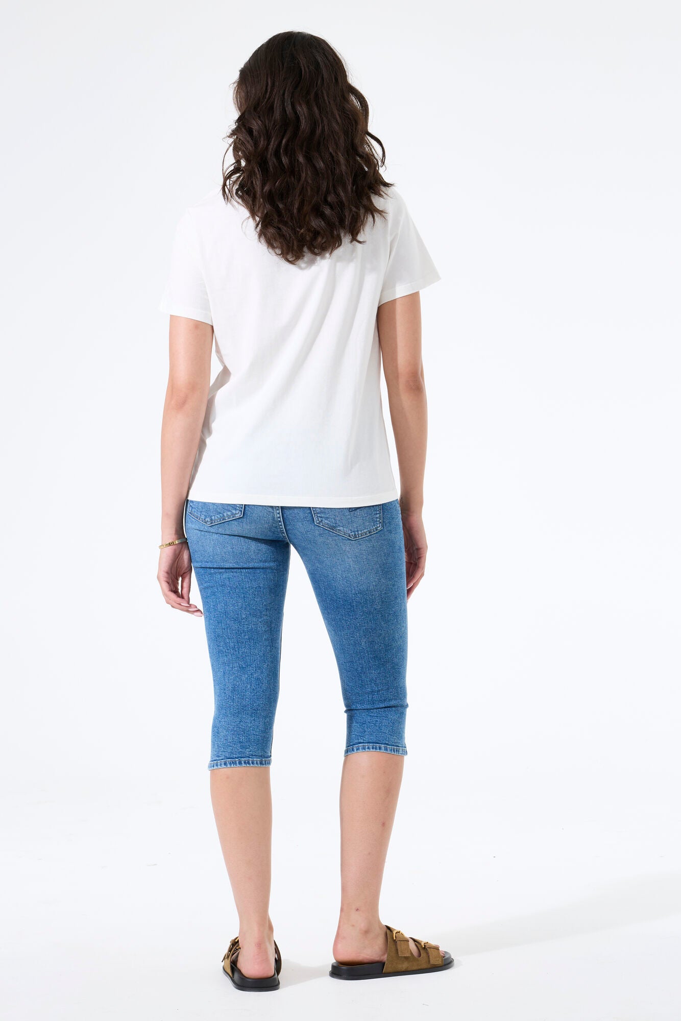 Back view of a woman wearing a slim-fit capri denim pant with a short-sleeve t-shirt and slide sandals.