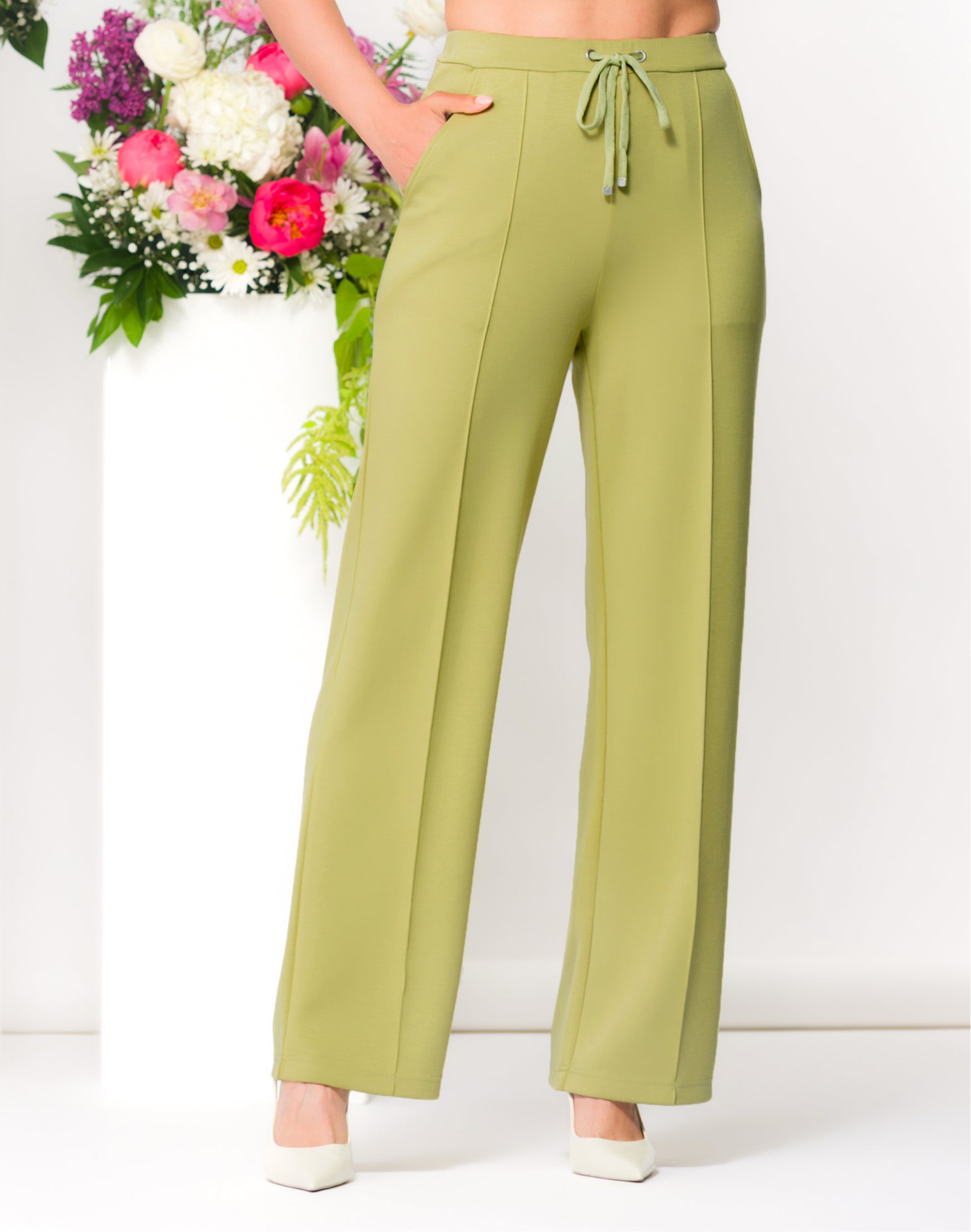 Woman with dark hair wearing a lime green wide-leg lounge pants with a drawstring waist, standing in a minimalist studio with floral arrangements behind her