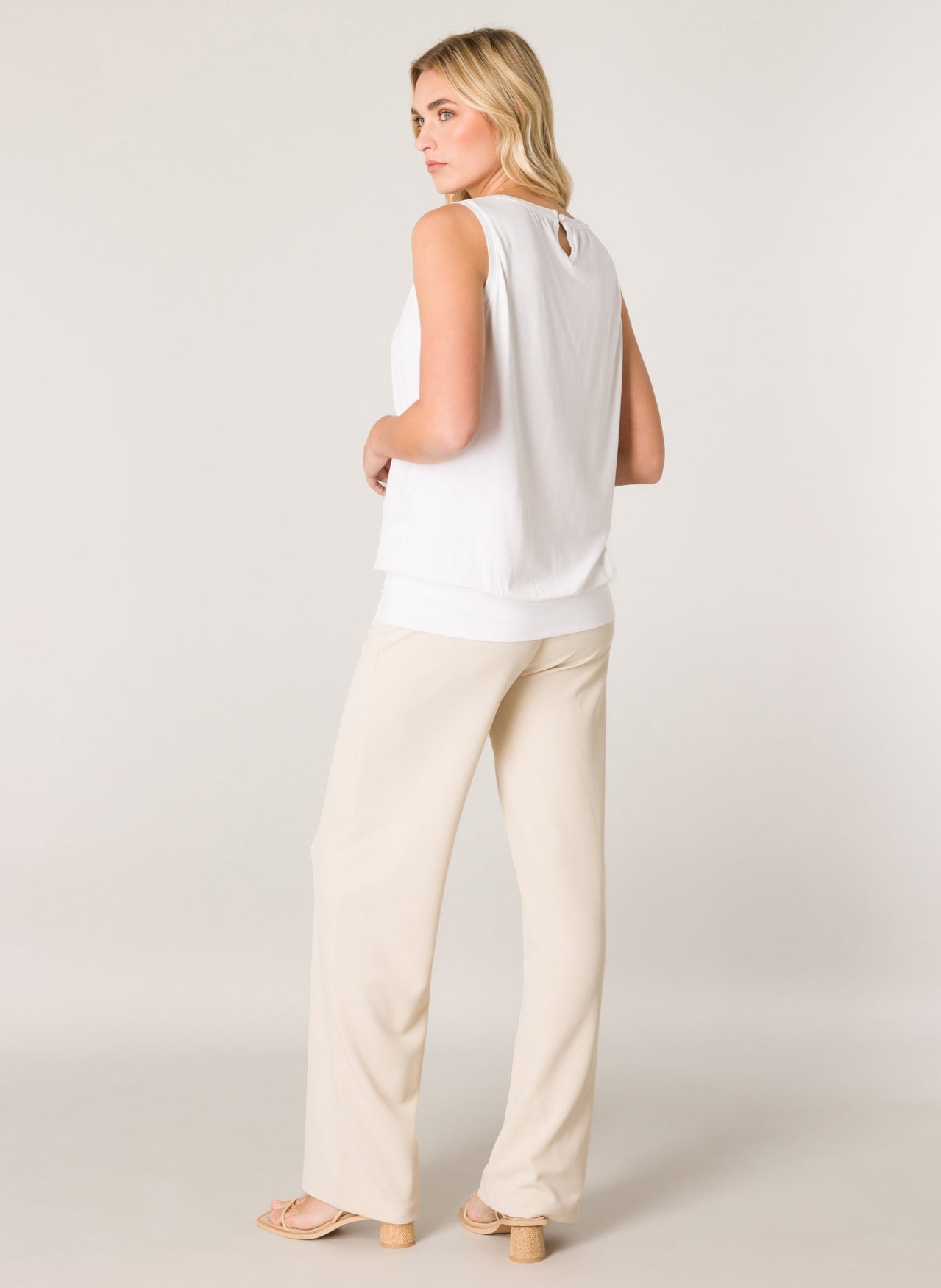 Women's white sleeveless blouse with a relaxed drape, wide waistband hem, and back keyhole detail, shown with cream wide-leg pants.