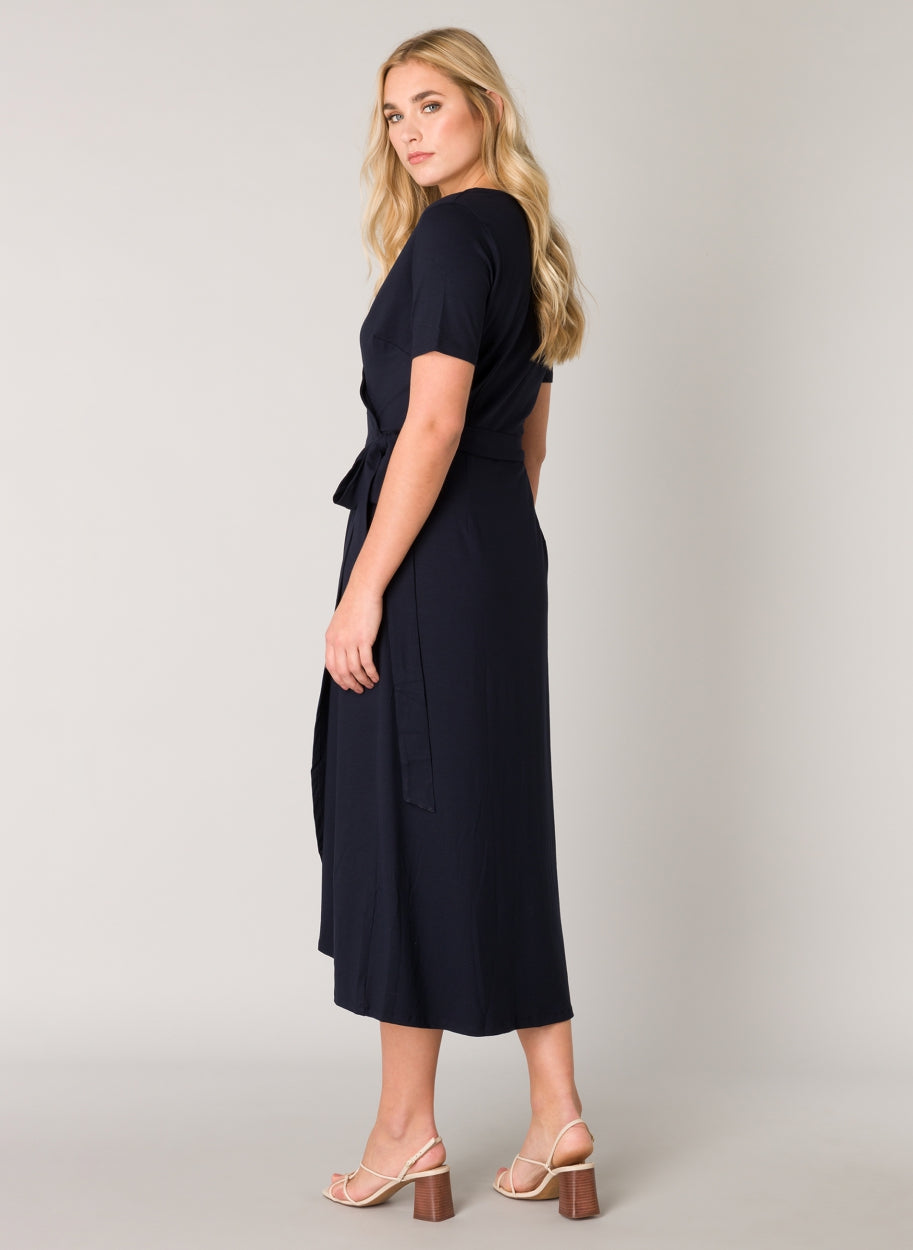 Women's dark blue wrap midi dress shown from the back, featuring short sleeves, a self-tie waist belt, and a flowing midi-length skirt. The smooth fabric offers an effortless drape and comfortable fit.