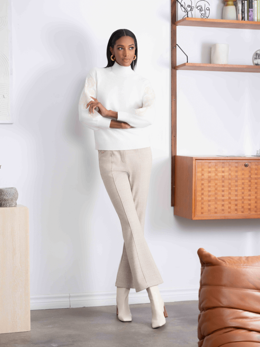 Woman in a white sweater and beige pants standing in a room with wooden shelves and a brown leather chair.