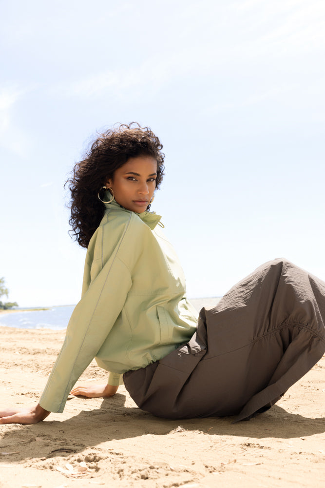 Model seated on warm sand by the shoreline, wearing a soft sage lightweight jacket paired with a relaxed olive midi skirt. Styled with gold hoop earrings and natural texture, the look evokes understated elegance and calm coastal sophistication under a bright, open sky.