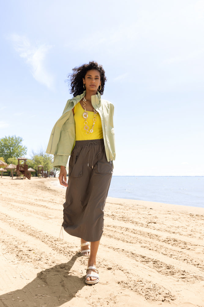 Model walking confidently along a sunlit beach, wearing a light sage zip-front jacket layered over a bright yellow cami and a flowing olive midi skirt. The look is finished with statement gold jewelry and flat sandals, capturing an effortless, modern coastal style against a serene shoreline backdrop.