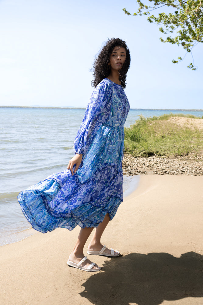 Woman wearing a blue abstract floral midi dress with long sleeves and tiered skirt, standing barefoot in white slide sandals on a sandy lakeside shore.