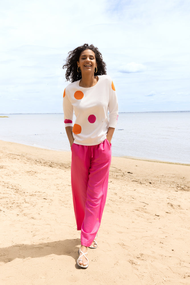 Woman walking on a sandy beach wearing a white sweater with orange and pink abstract polka dots, bright pink wide-leg pants, and white sandals.