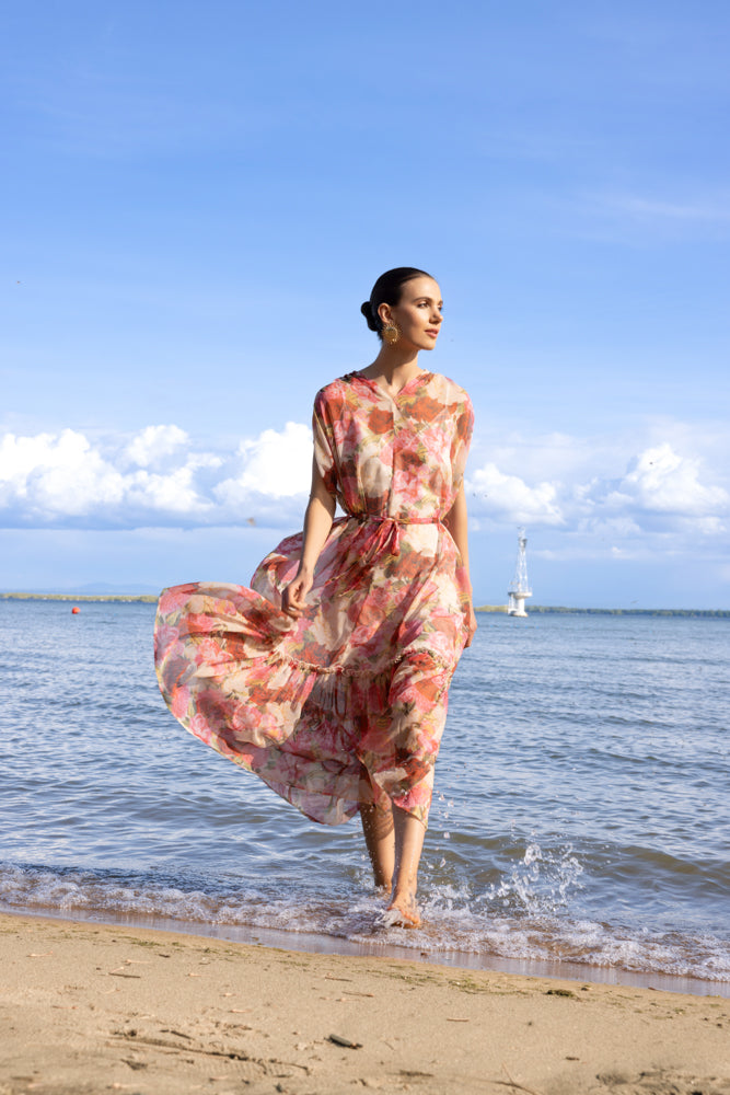 Model in a pink floral midi dress with cap sleeves and adjustable waist tie, standing by the water on a sunny day.