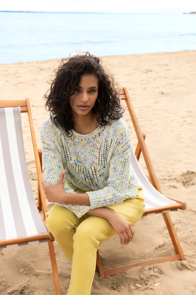 Model wearing a blue and yellow melange lightweight sweater with airy pointelle knit detail and relaxed fit, paired with yellow trousers, sitting on a sandy beach by the water.