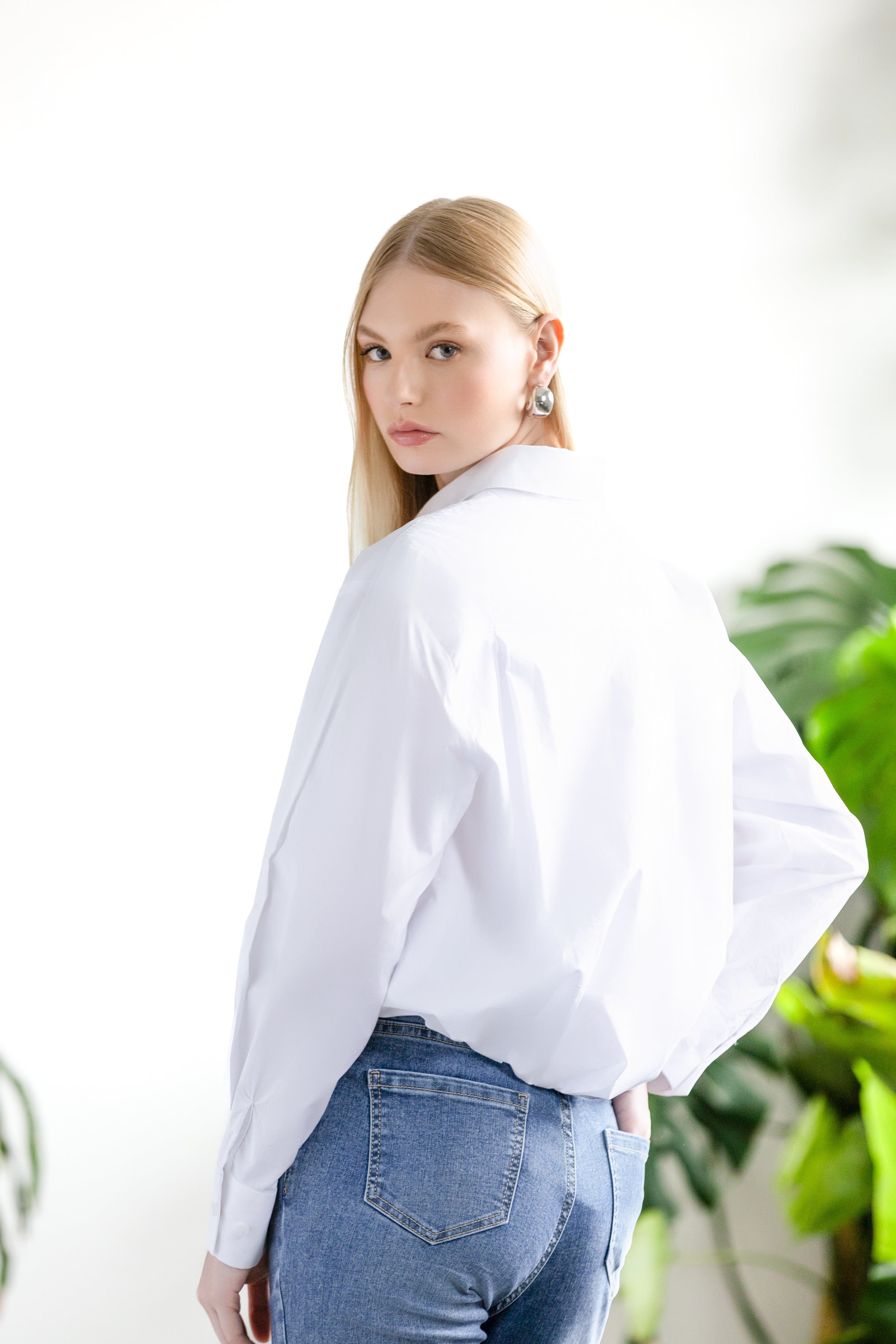 Woman with light hair wearing a white blouse and light jeans in a white studio surrounded by green leafy plants.