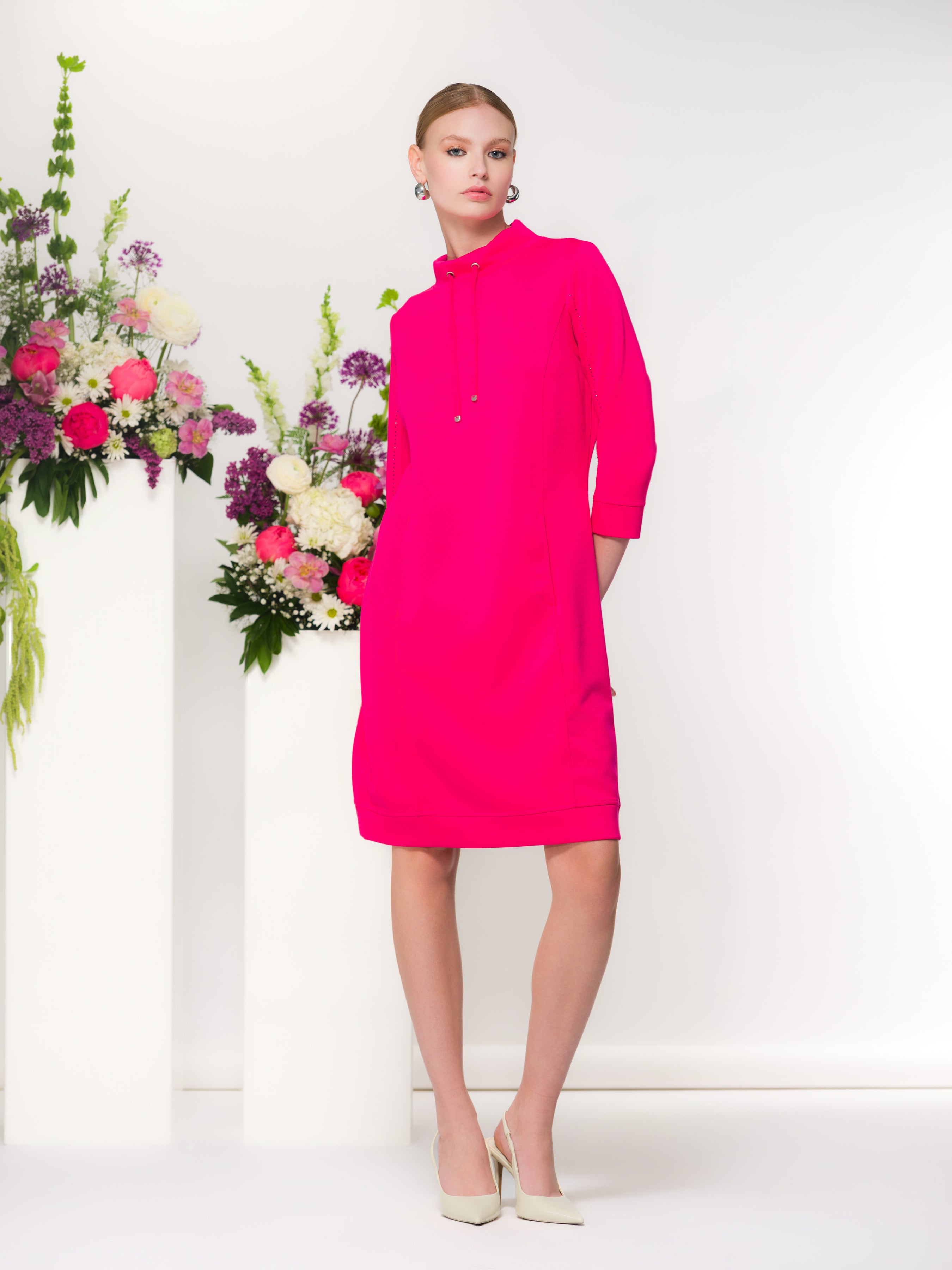 Woman wearing a bright pink dress standing in front of floral decorations.