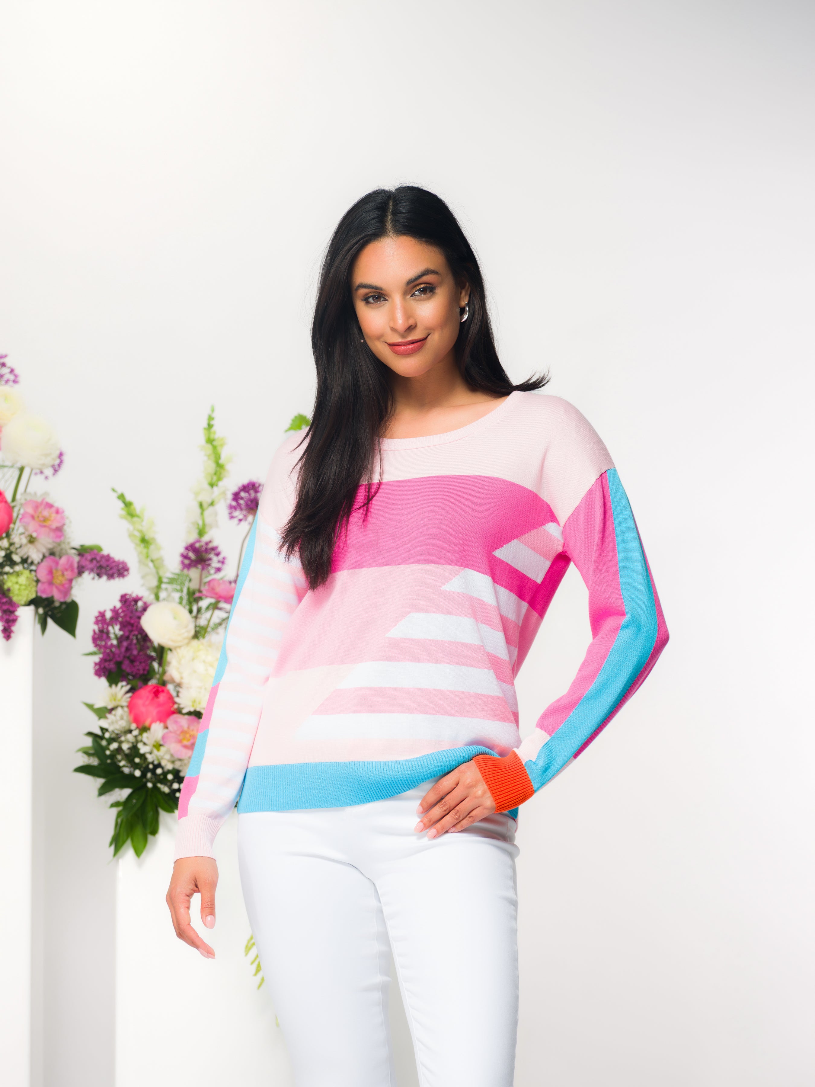 Model wearing a pastel pink geometric print sweater with blue accents and white pants standing in front of a floral arrangement.
