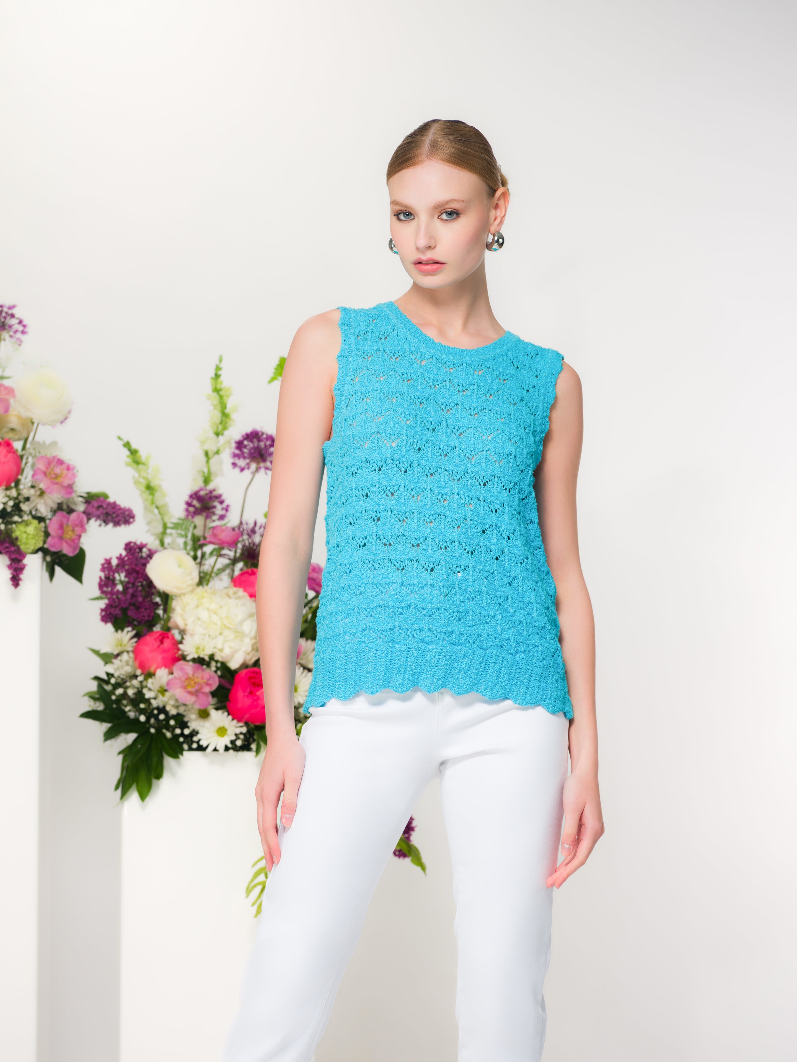 Woman wearing a sleeveless textured turquoise knit top with a crew neckline and scalloped hem, paired with fitted pants, standing in a bright studio setting with floral accents in the background.
