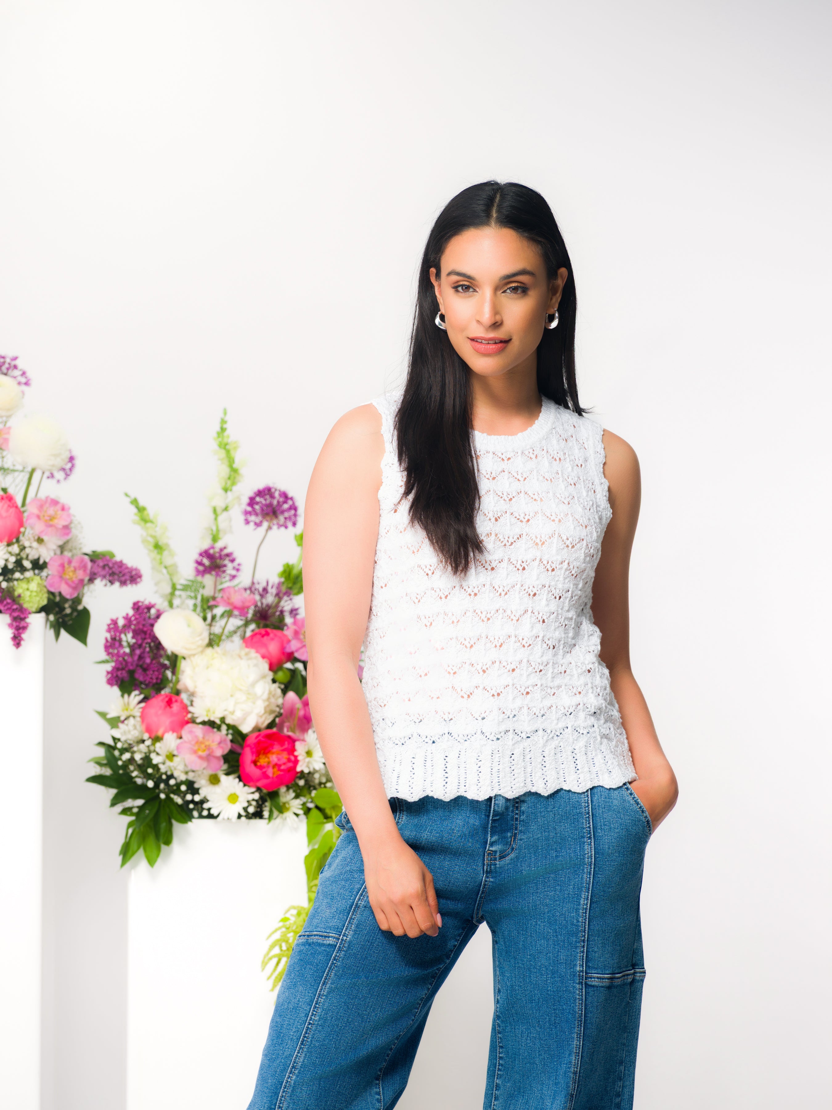 Woman wearing a sleeveless textured white knit top with a crew neckline and scalloped hem, paired with fitted pants, standing in a bright studio setting with floral accents in the background.