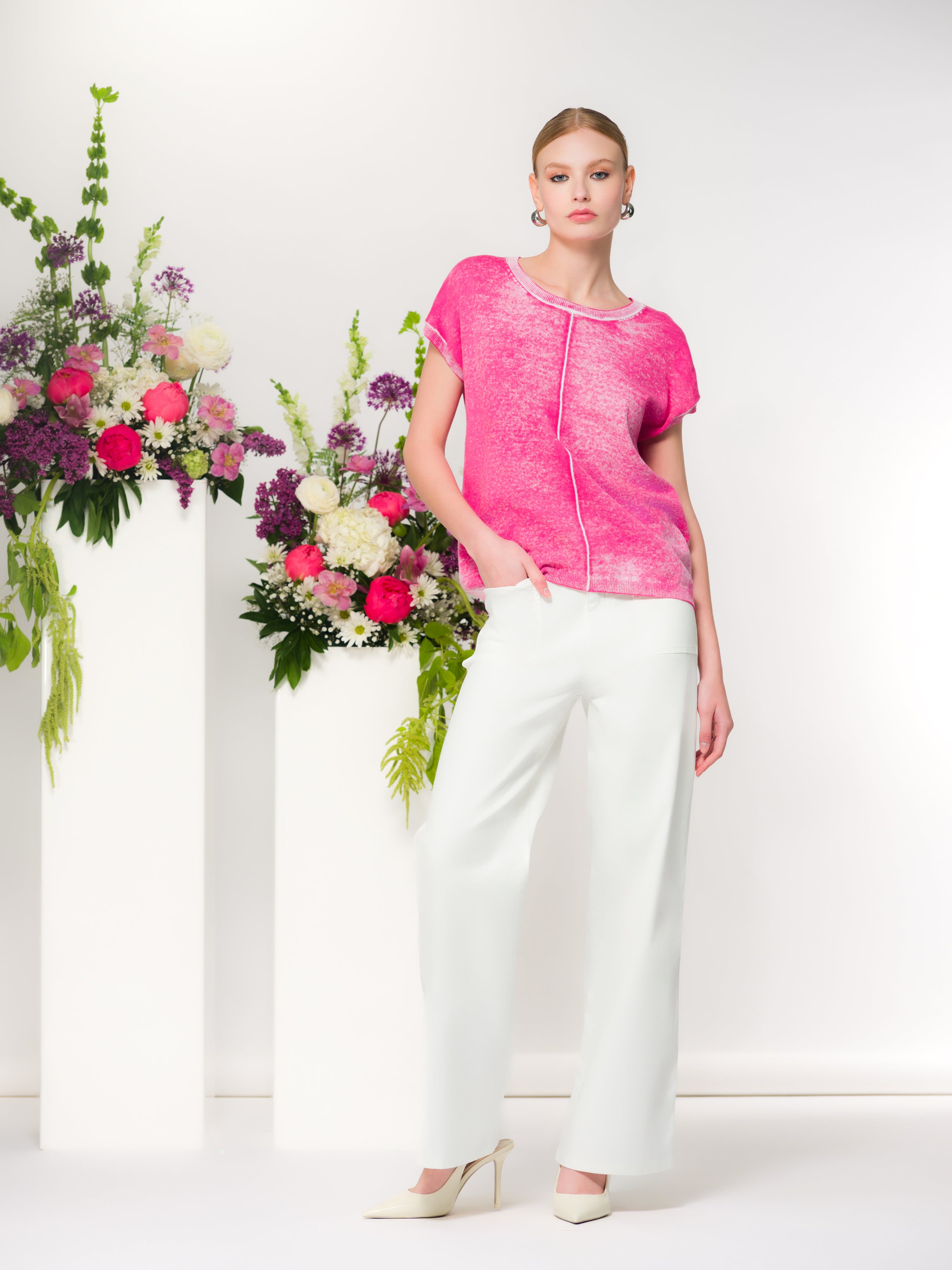 Woman wearing a fuchsia short-sleeve knit top with a relaxed silhouette and soft drape, styled with wide-leg pants and heels. She stands in a bright studio with decorative floral arrangements in the background.
