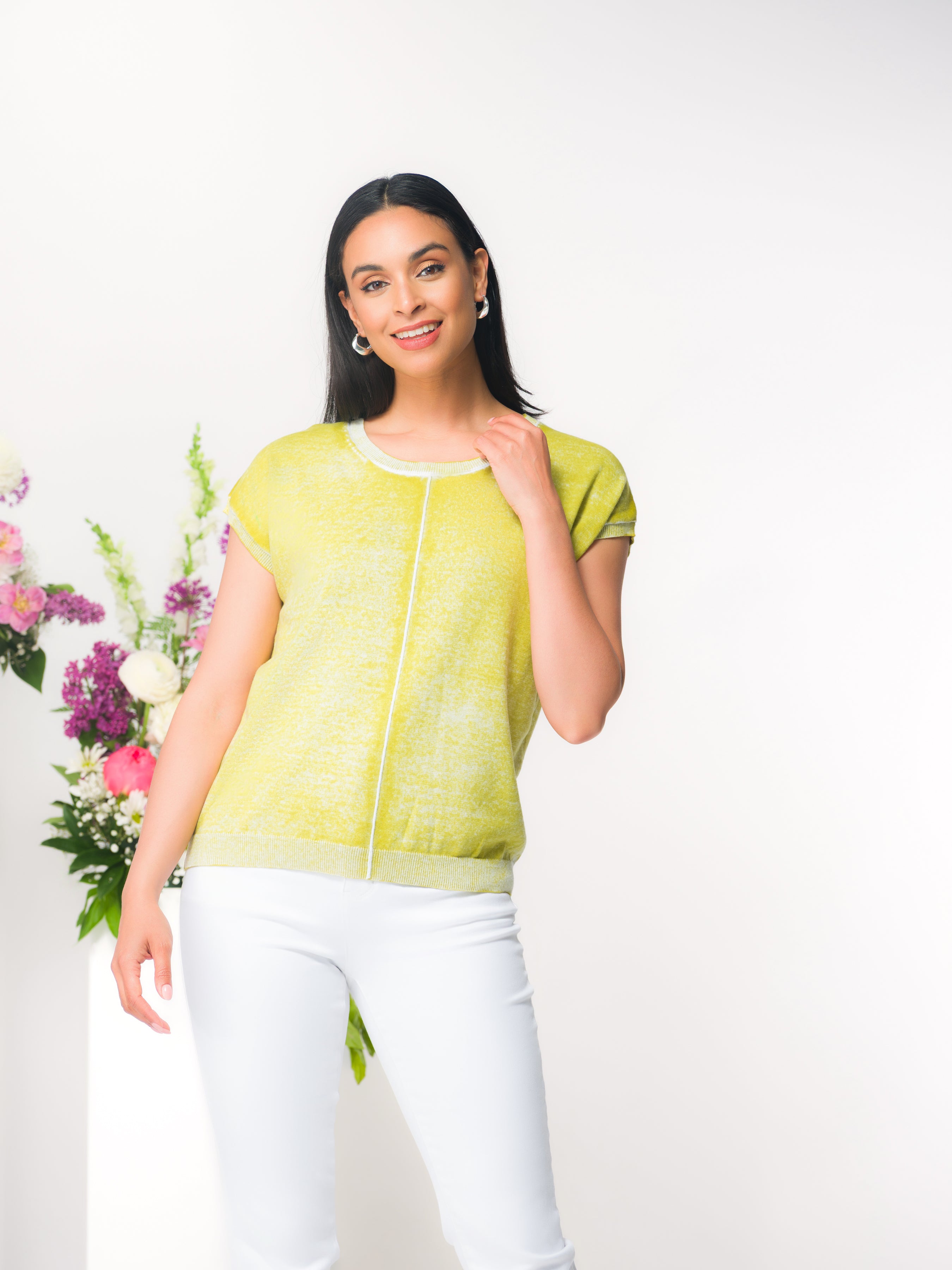 Woman wearing a lime short-sleeve knit top with a relaxed fit and subtle front seam detail, paired with straight-leg pants. She is standing in a studio setting with floral arrangements on white plinths.