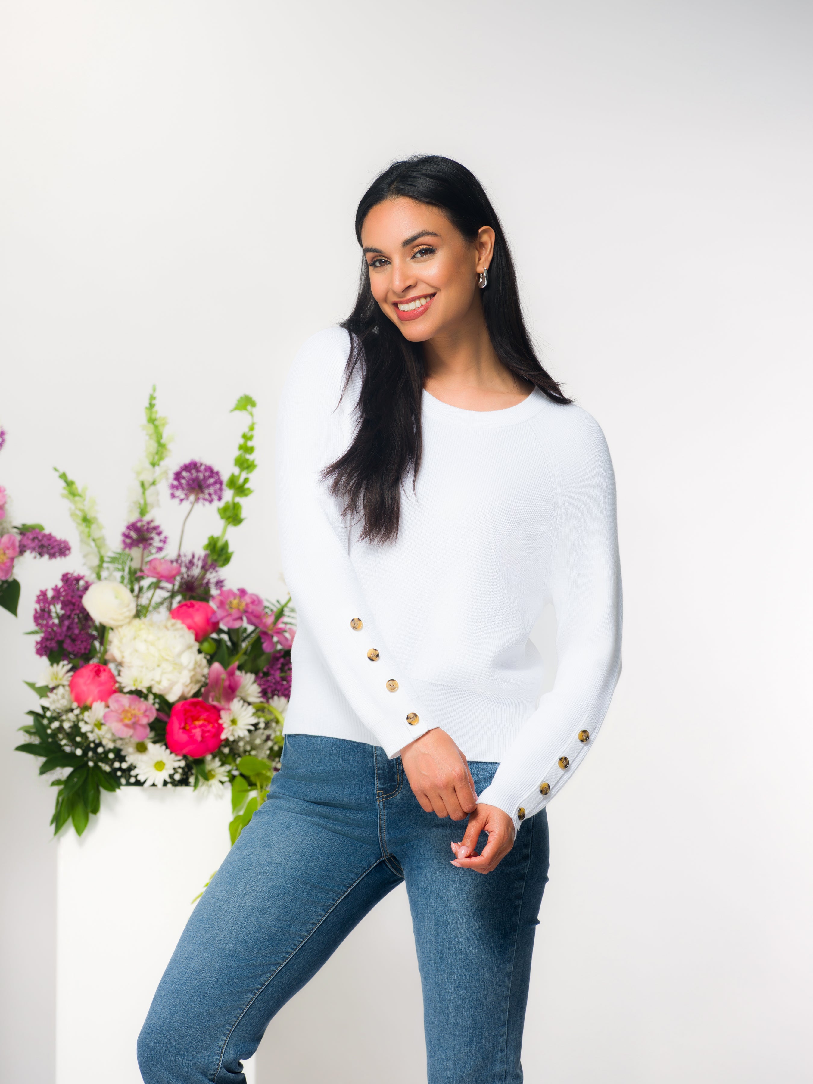 Woman wearing a white sweater with jeans standing next to a floral arrangement with a white background