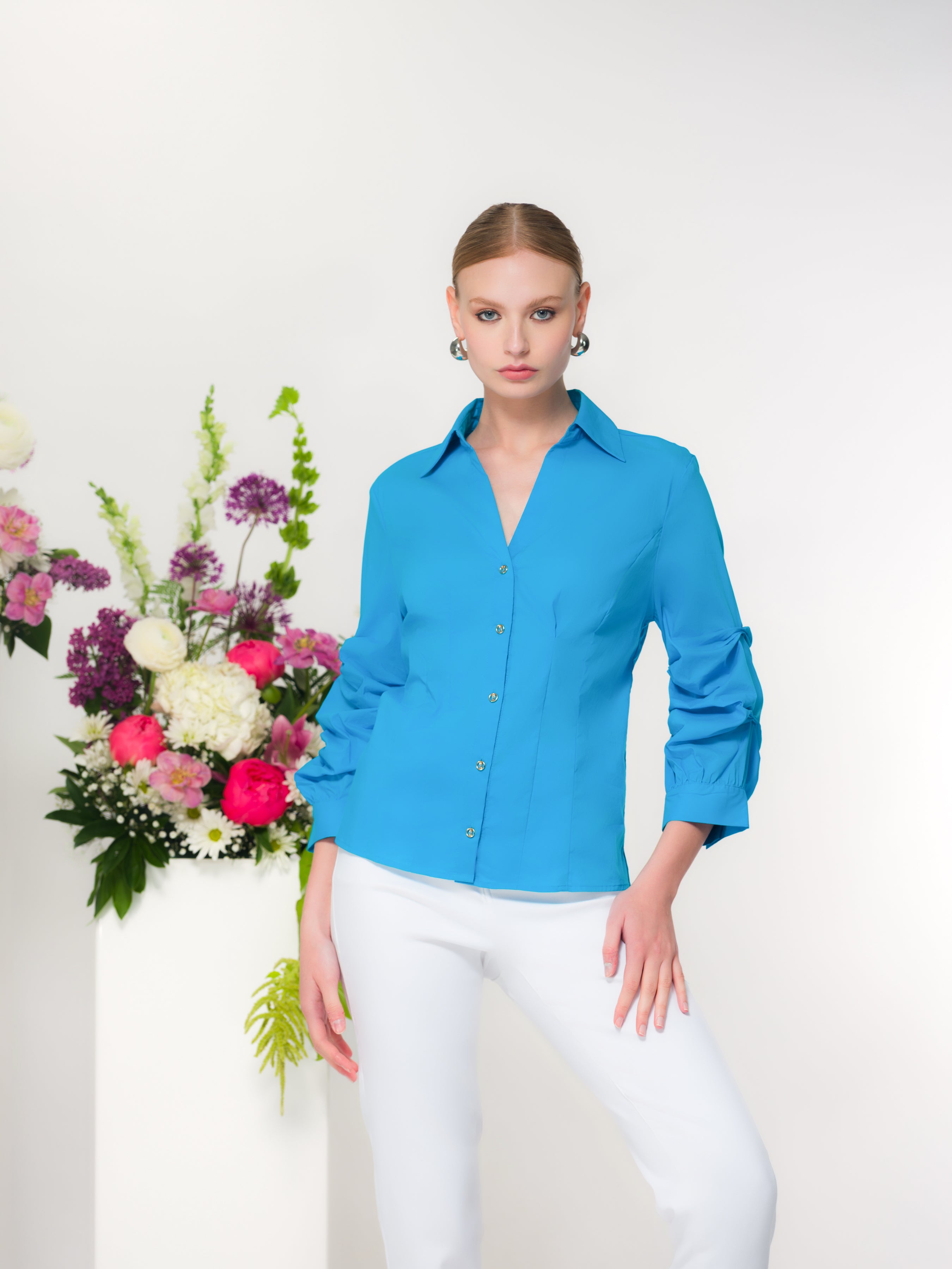 Woman with blonde hair wearing a turquoise blouse with white pants standing in front of a floral arrangement with a white background