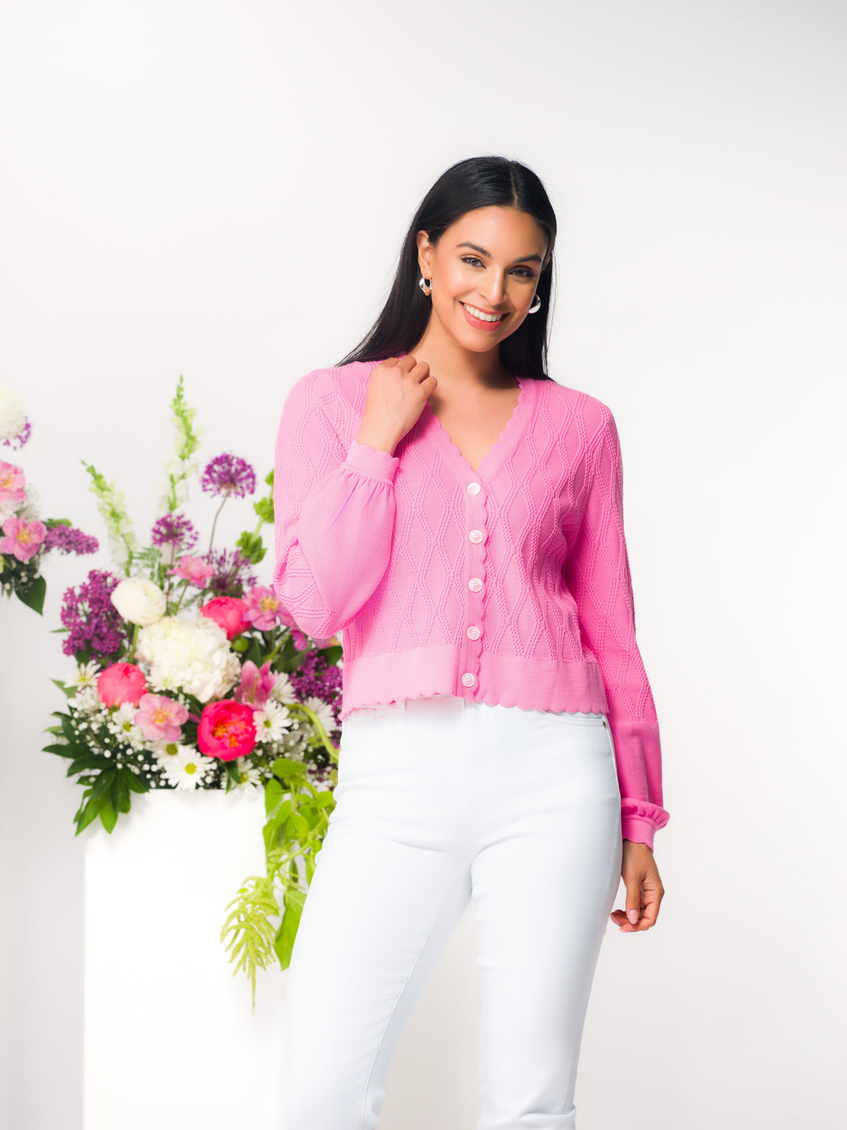 Model wearing a pink fitted button-front knit cardigan with scalloped neckline and long sleeves, styled with white pants, photographed in a studio setting.