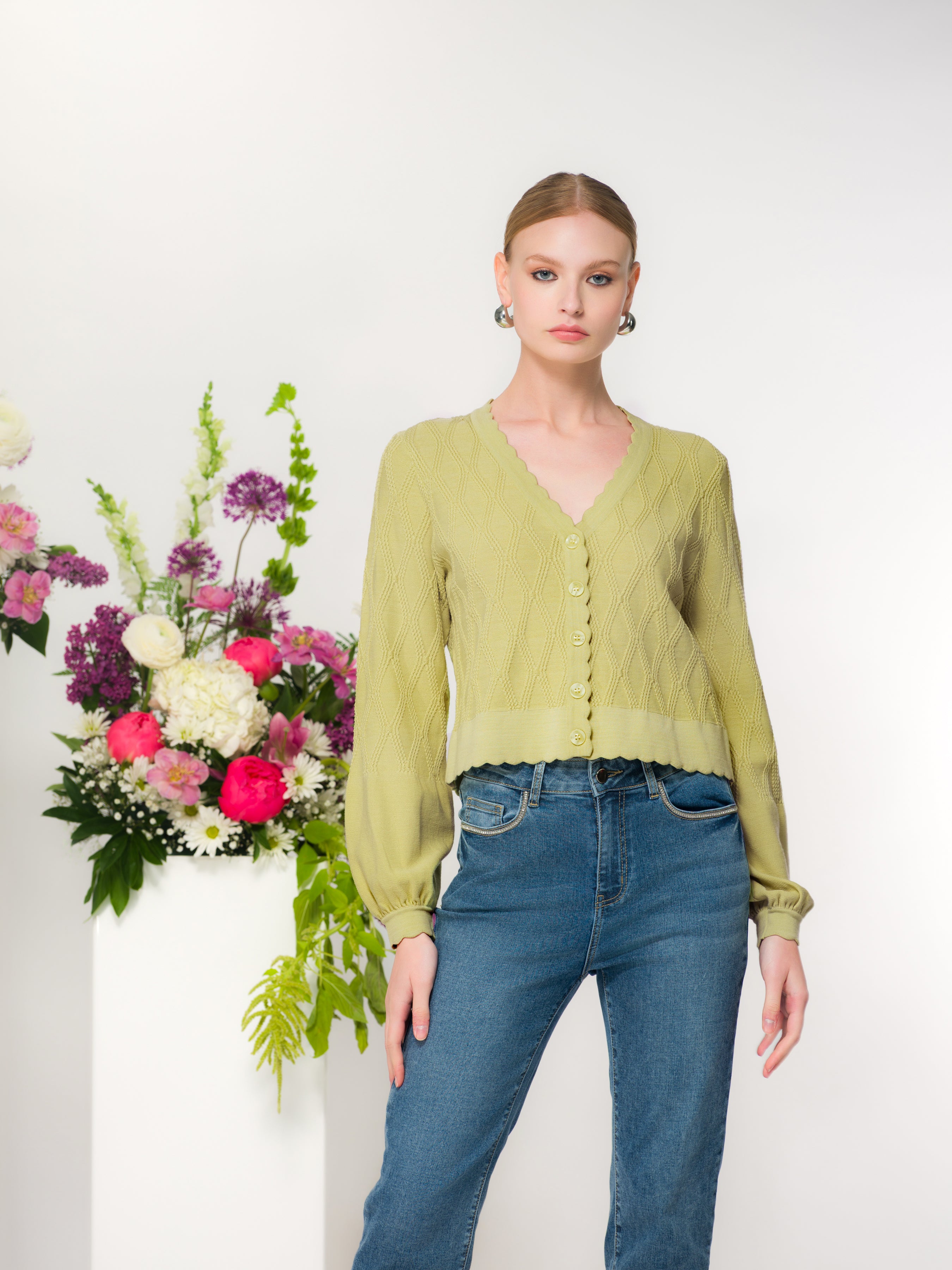 Model wearing a green fitted button-front knit cardigan with scalloped neckline and long sleeves, styled with denim jeans, photographed in a studio setting.