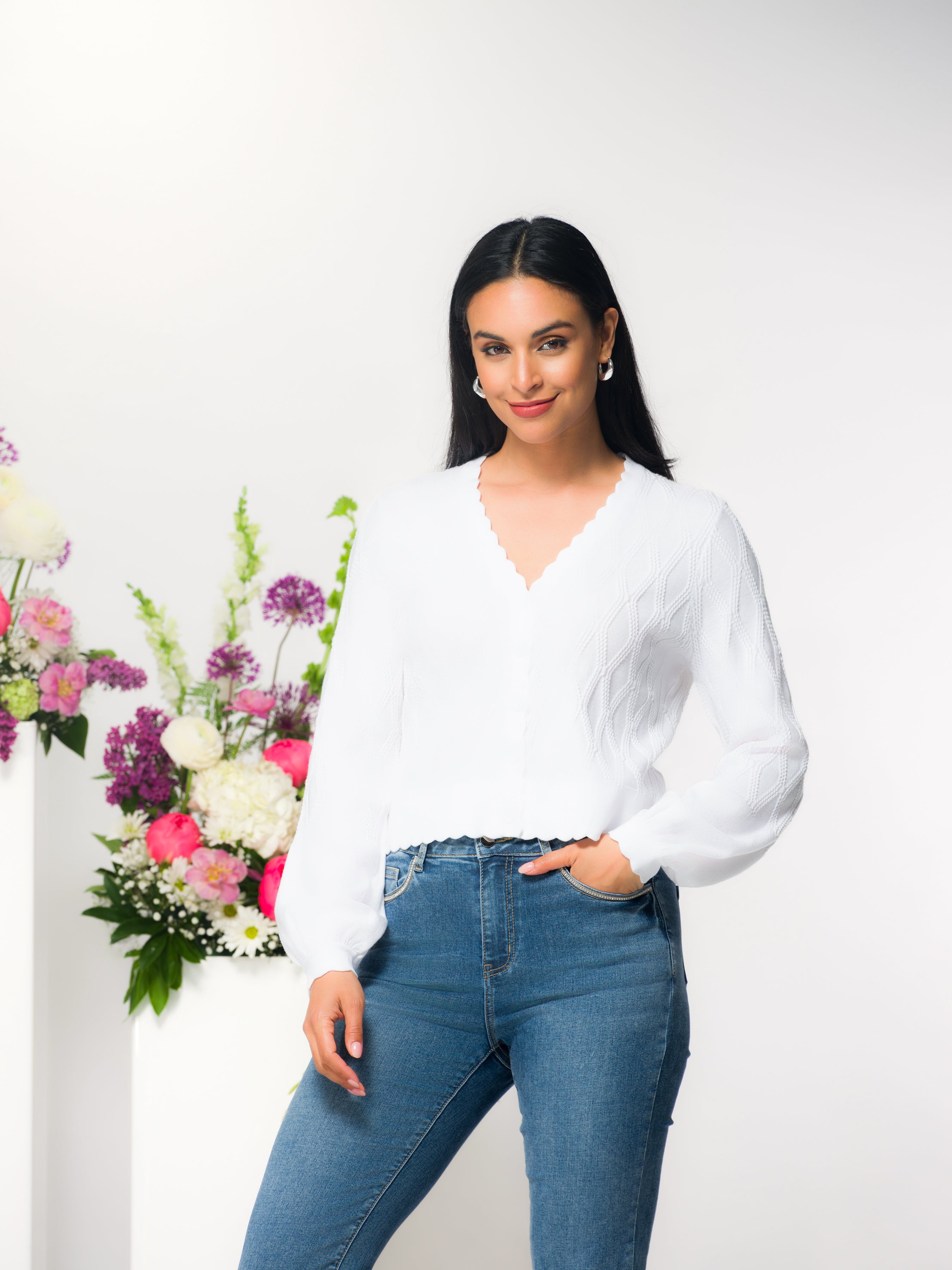 Model wearing a white fitted button-front knit cardigan with scalloped neckline and long sleeves, styled with denim jeans, photographed in a studio setting.