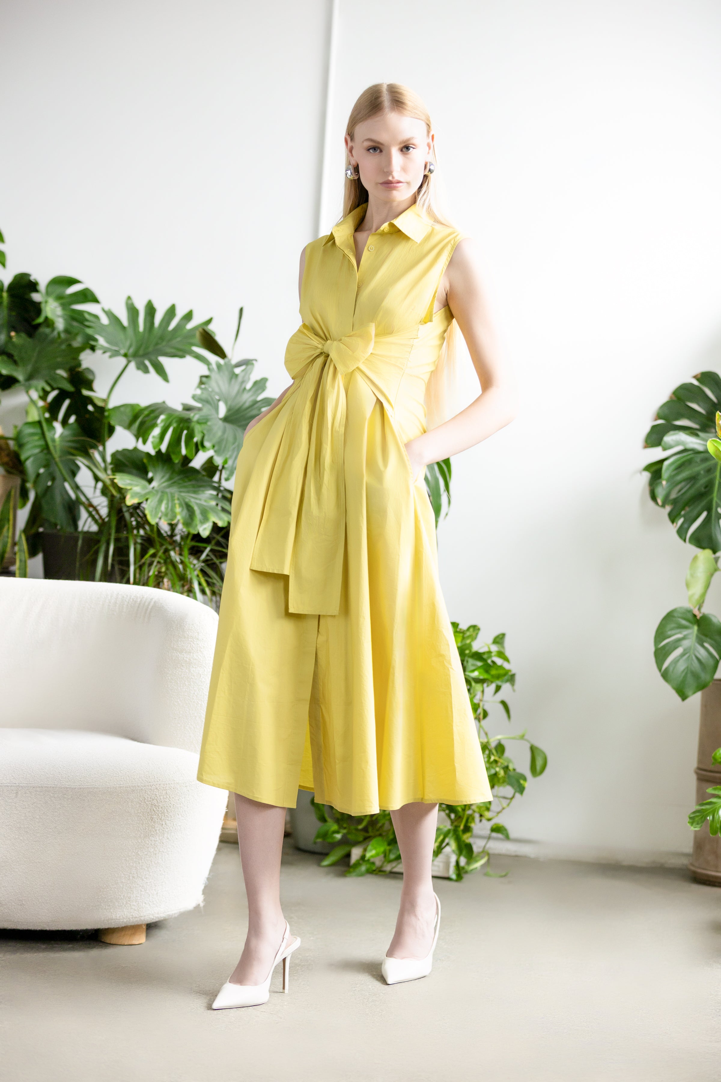 Model wearing a lime green sleeveless midi dress with a wrap-style waist and collared neckline, featuring a softly draped skirt and side pockets, styled with classic heels in a studio setting with greenery.