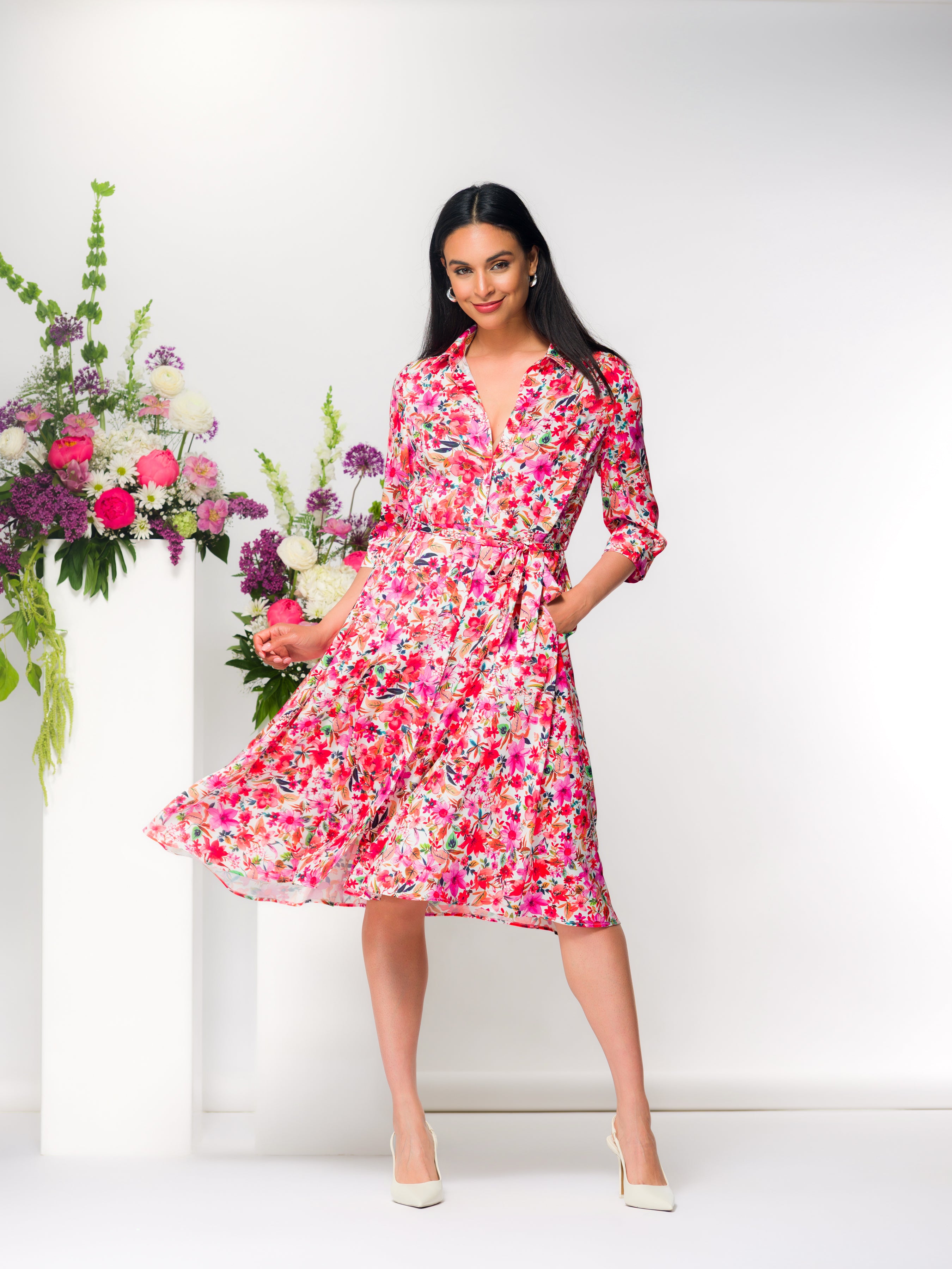 Woman wearing a button-front shirt dress with a waist tie and three-quarter length sleeves, styled indoors with floral arrangements.