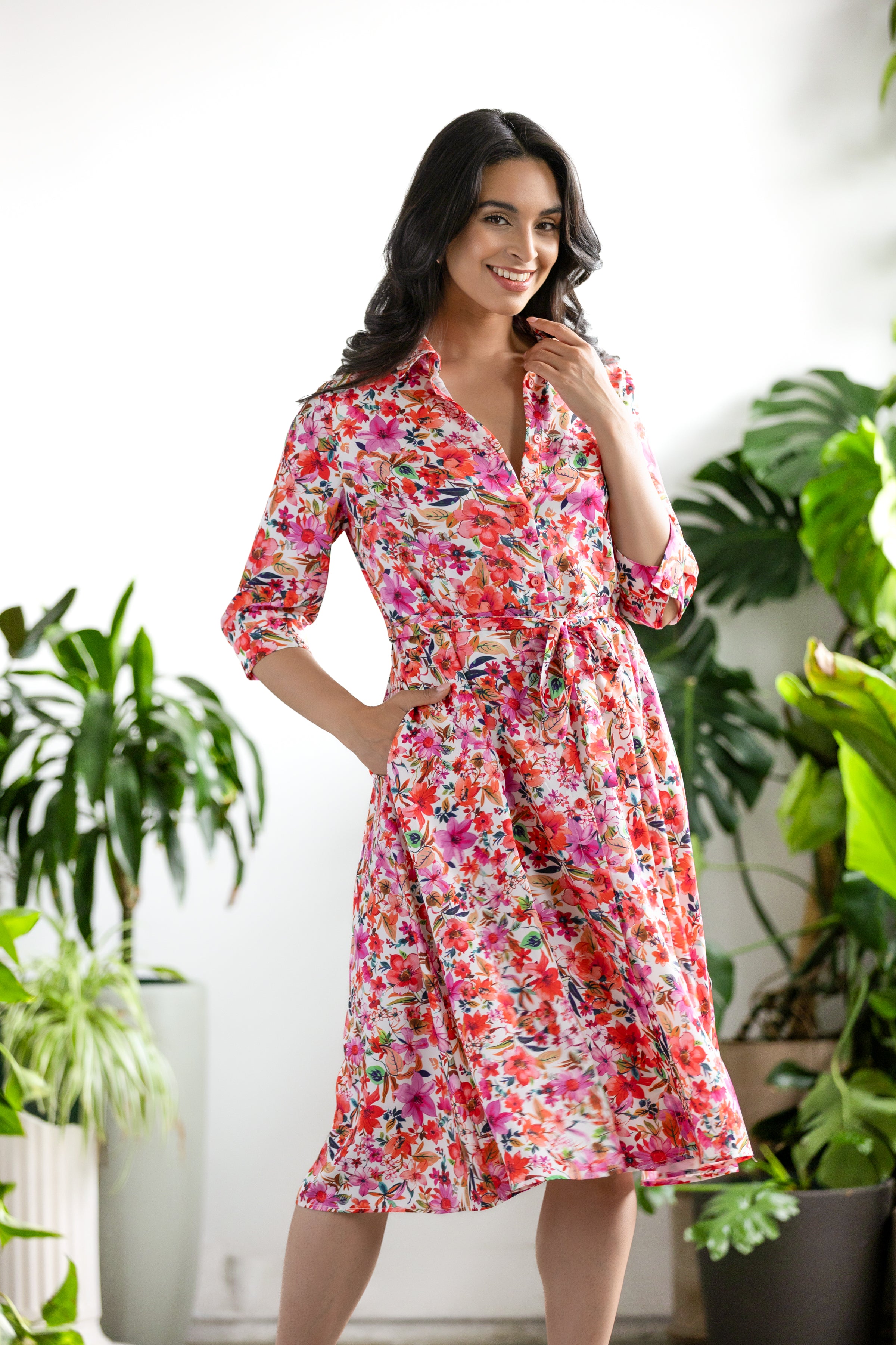 Woman wearing a button-front shirt dress with a waist tie and three-quarter length sleeves, styled indoors with plants.