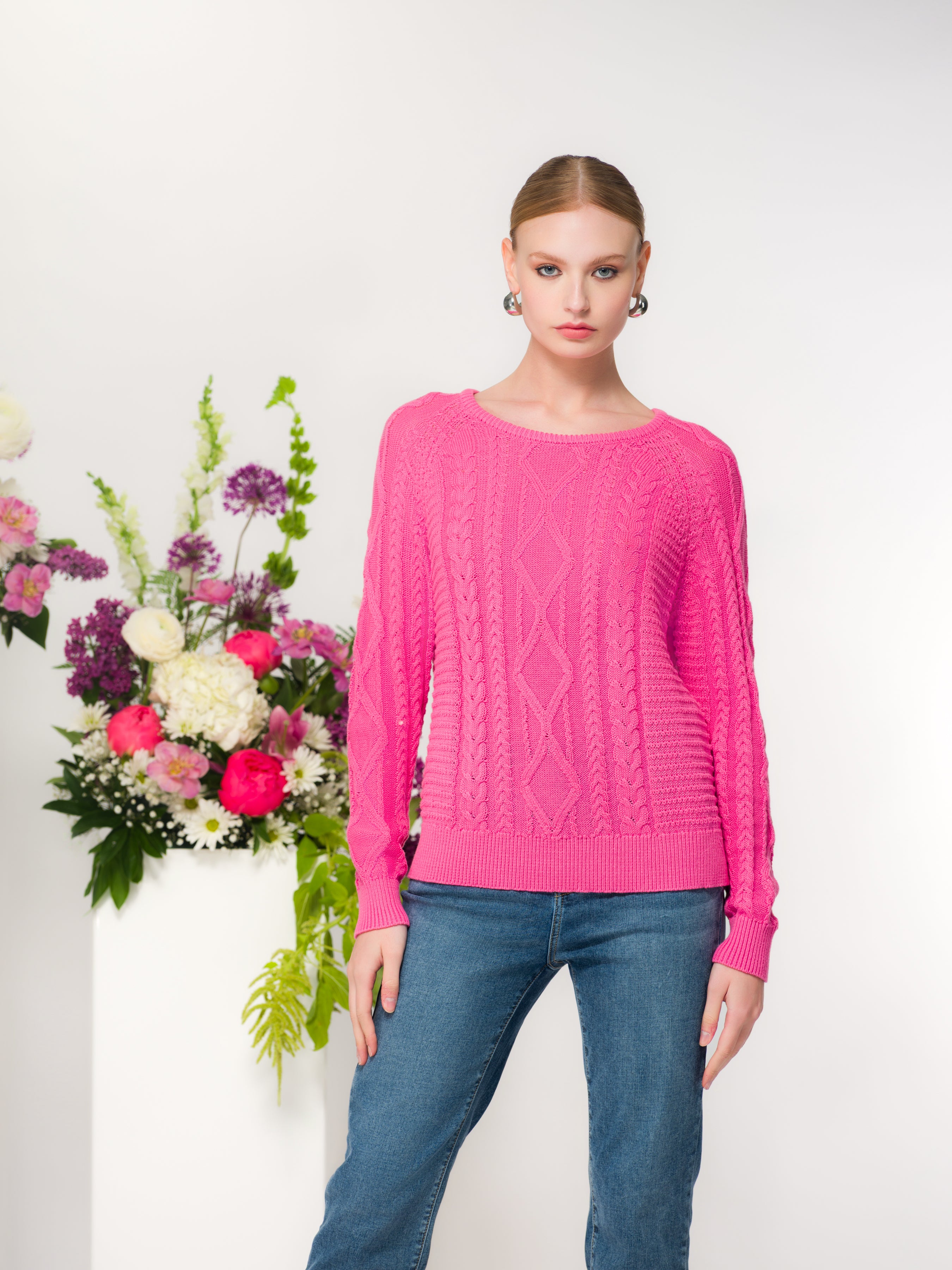 Model wearing a pink cable knit pullover sweater with a crew neckline and long sleeves, styled with jeans in a studio setting.