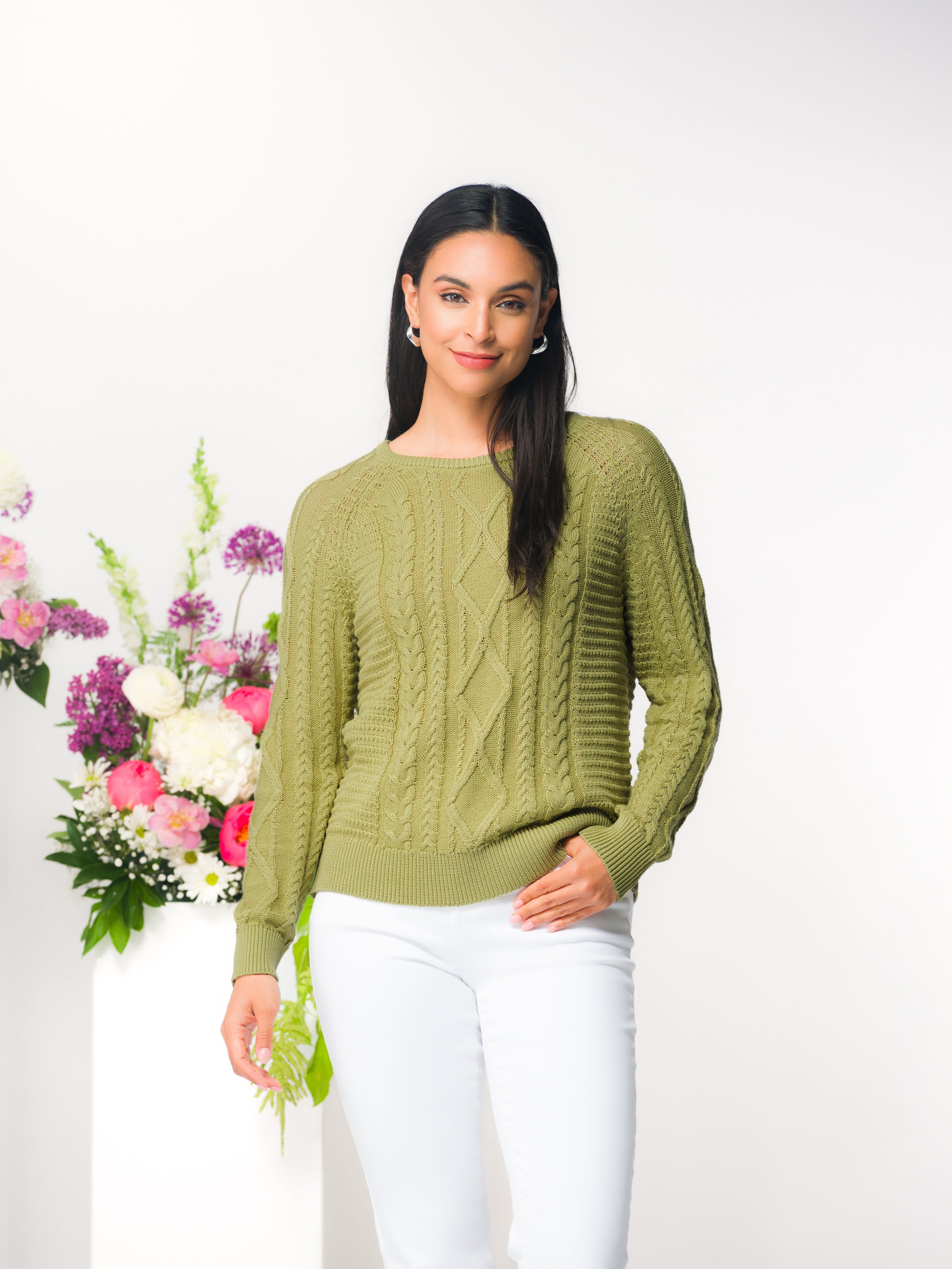Model wearing a green cable knit pullover sweater with a crew neckline and long sleeves, styled with white pants in a studio setting.