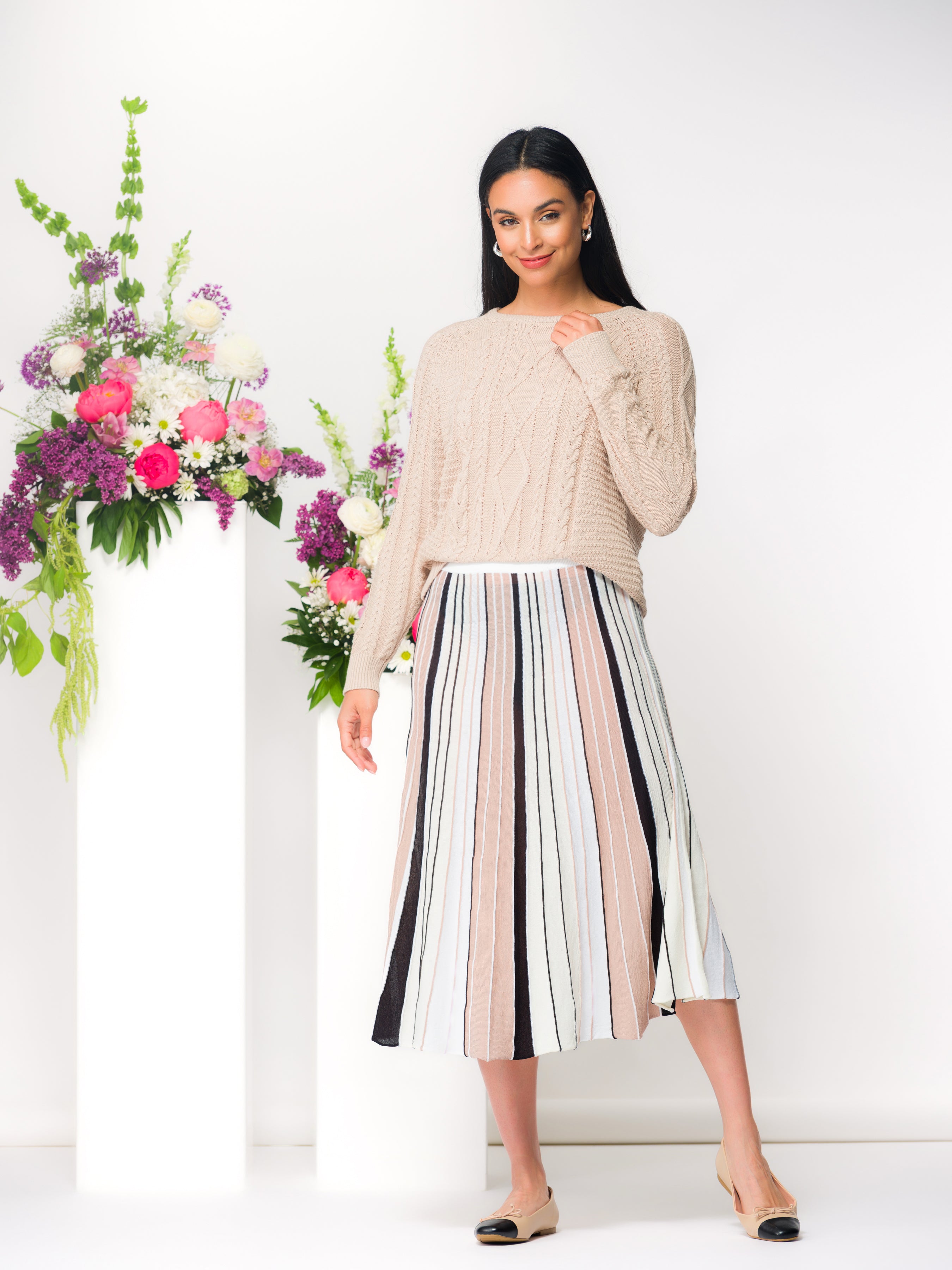 Model wearing a sand cable knit pullover sweater with a crew neckline and long sleeves, styled with a striped skirt in a studio setting.