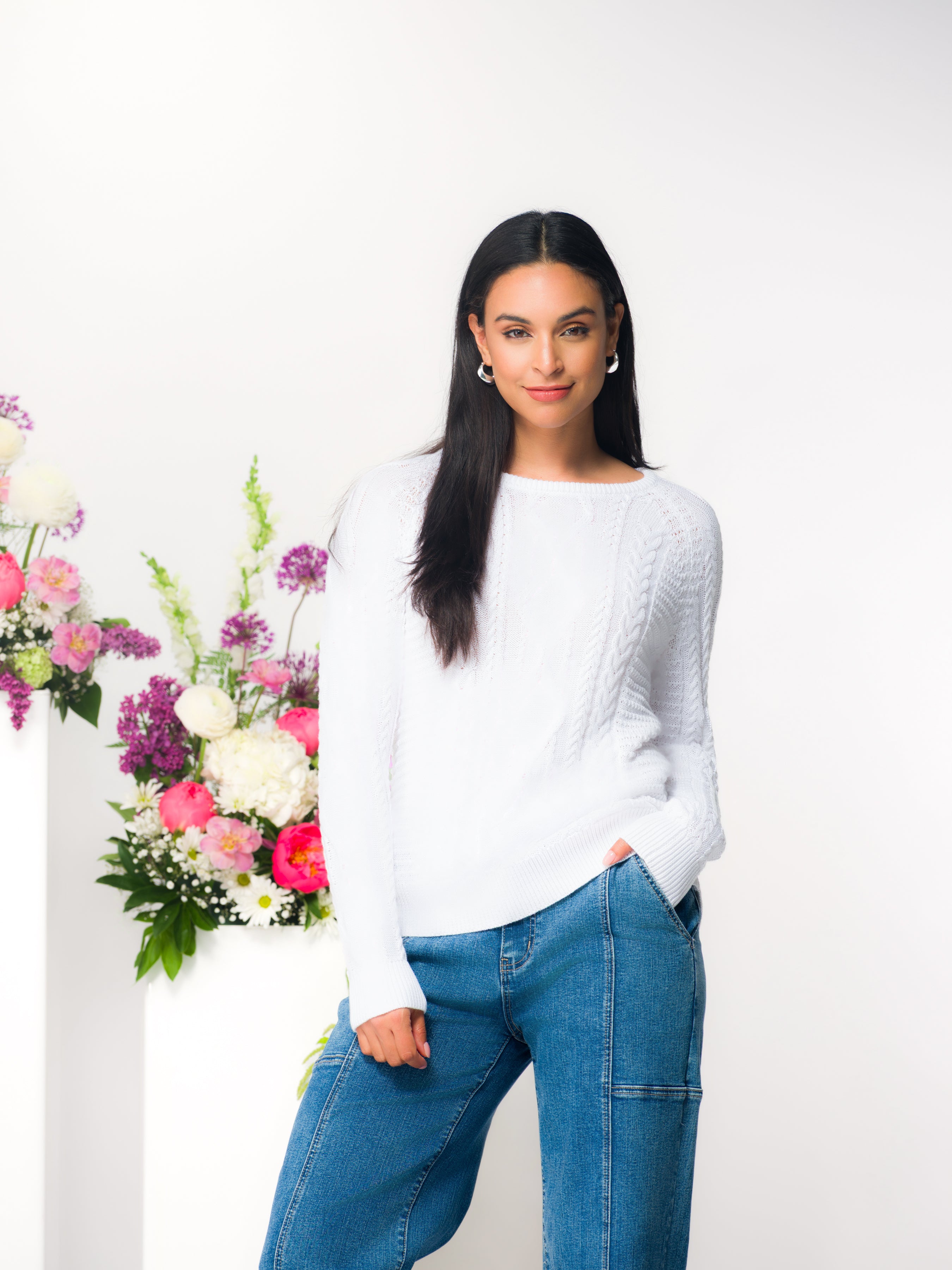 Model wearing a white cable knit pullover sweater with a crew neckline and long sleeves, styled with jeans in a studio setting.