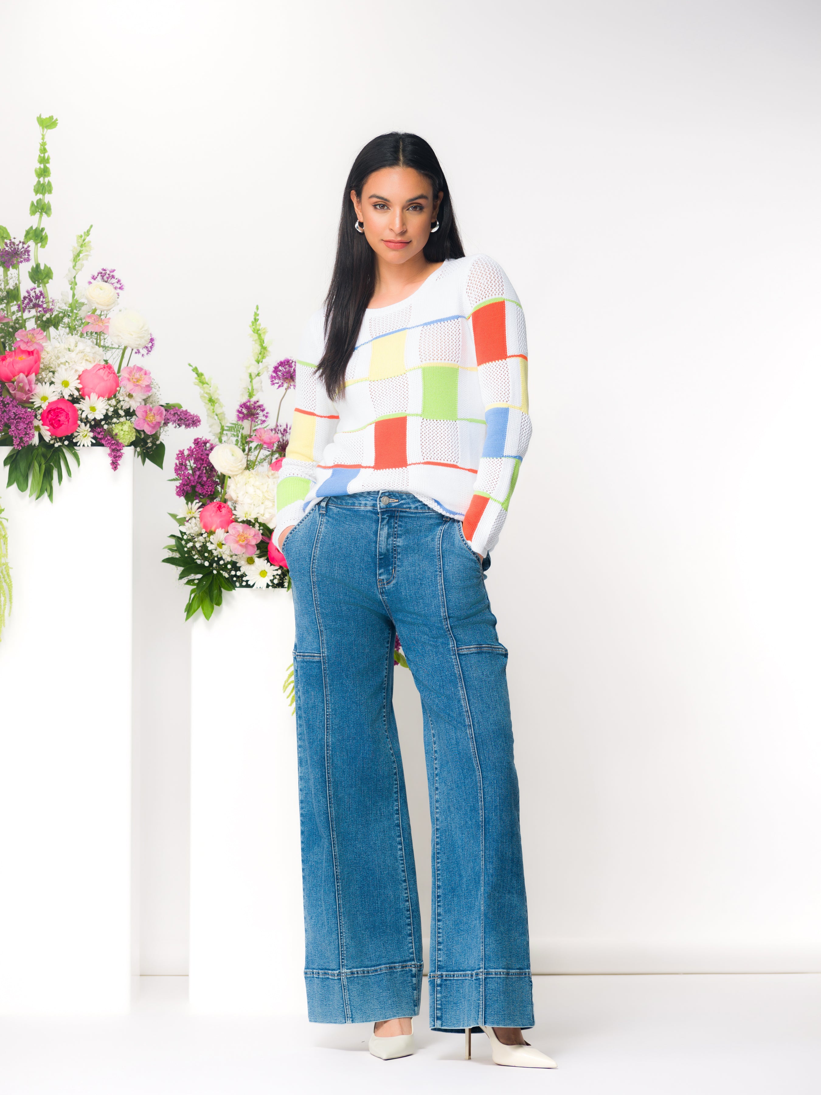 Woman wearing a long-sleeve cotton knit sweater with a textured geometric pattern, styled with wide-leg denim jeans and heels, standing in a studio setting with floral arrangements.