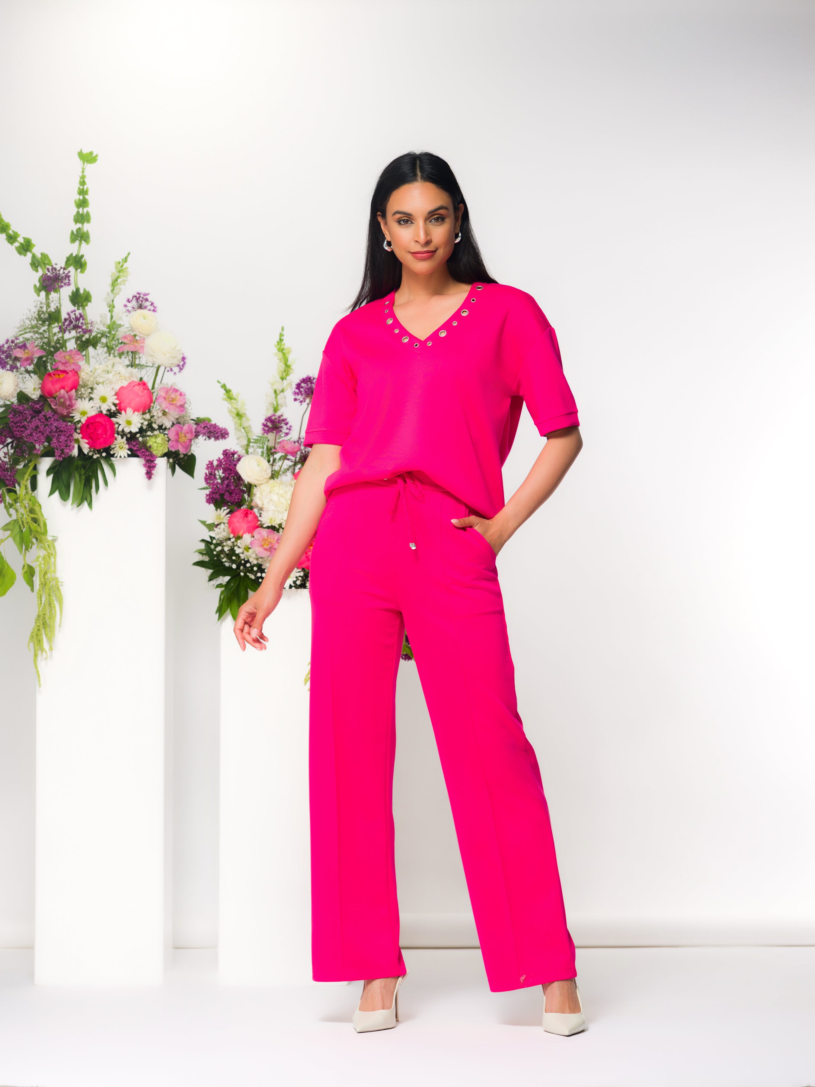 Woman wearing a relaxed pink V-neck top with short sleeves and matching wide-leg pants, styled indoors with floral arrangements.