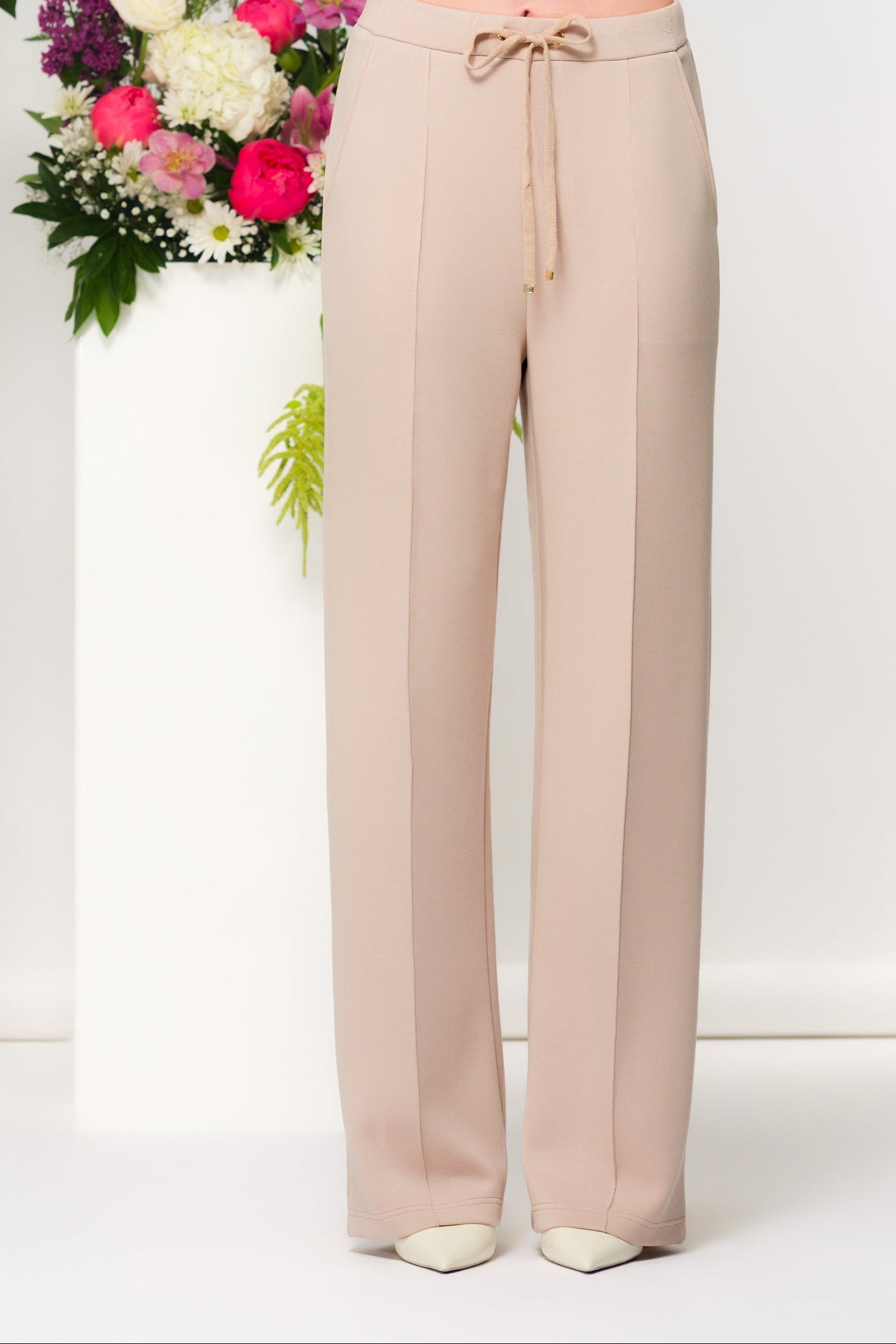 Woman with blonde wearing sand coloured wide-leg lounge pants with a drawstring waist, standing in a minimalist studio with floral arrangements behind her