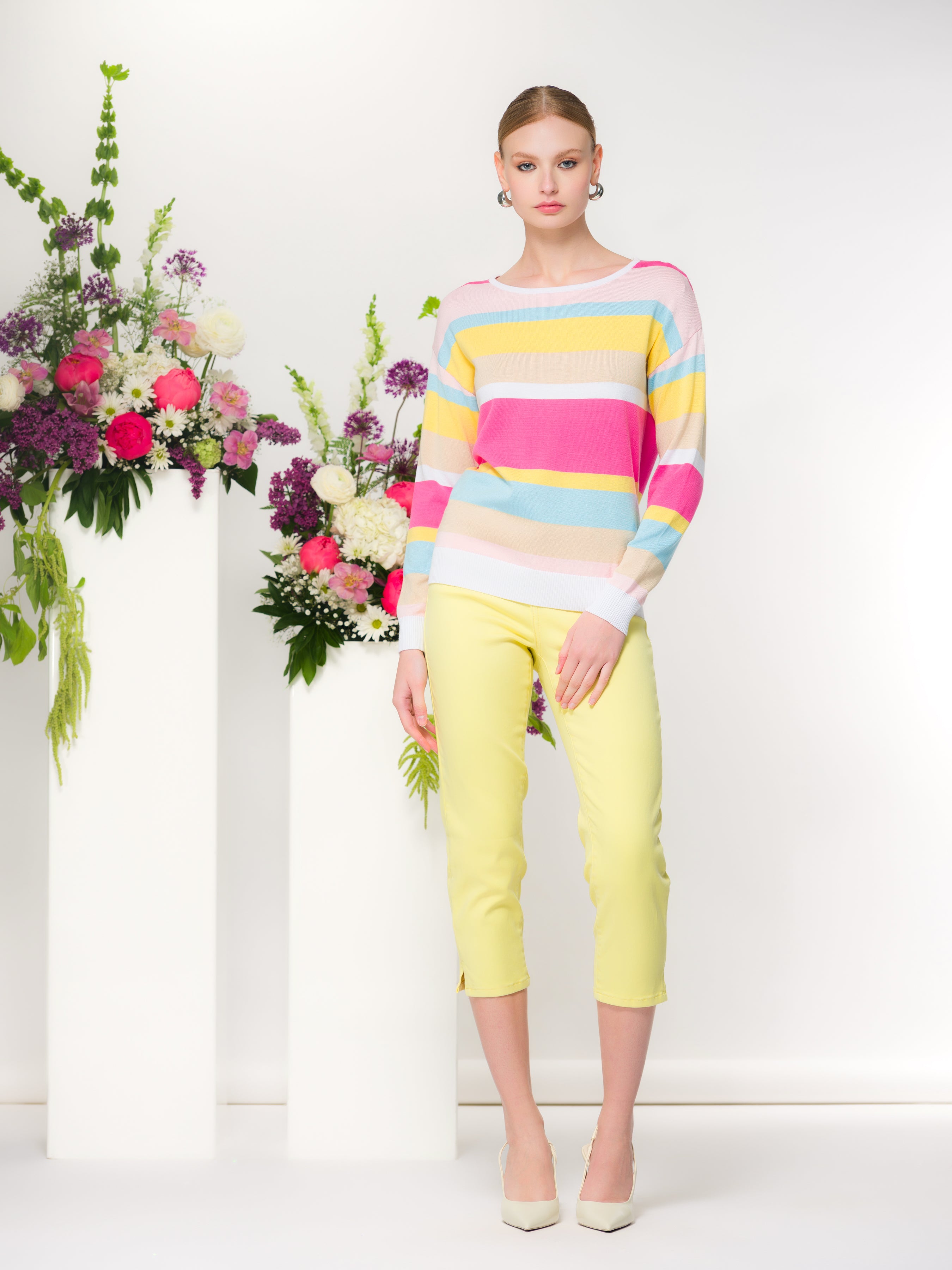 A studio photo of a women’s knit pullover with long sleeves and a relaxed fit, styled with cropped trousers and heels, shown on a model standing between minimalist floral arrangements against a clean, light background.