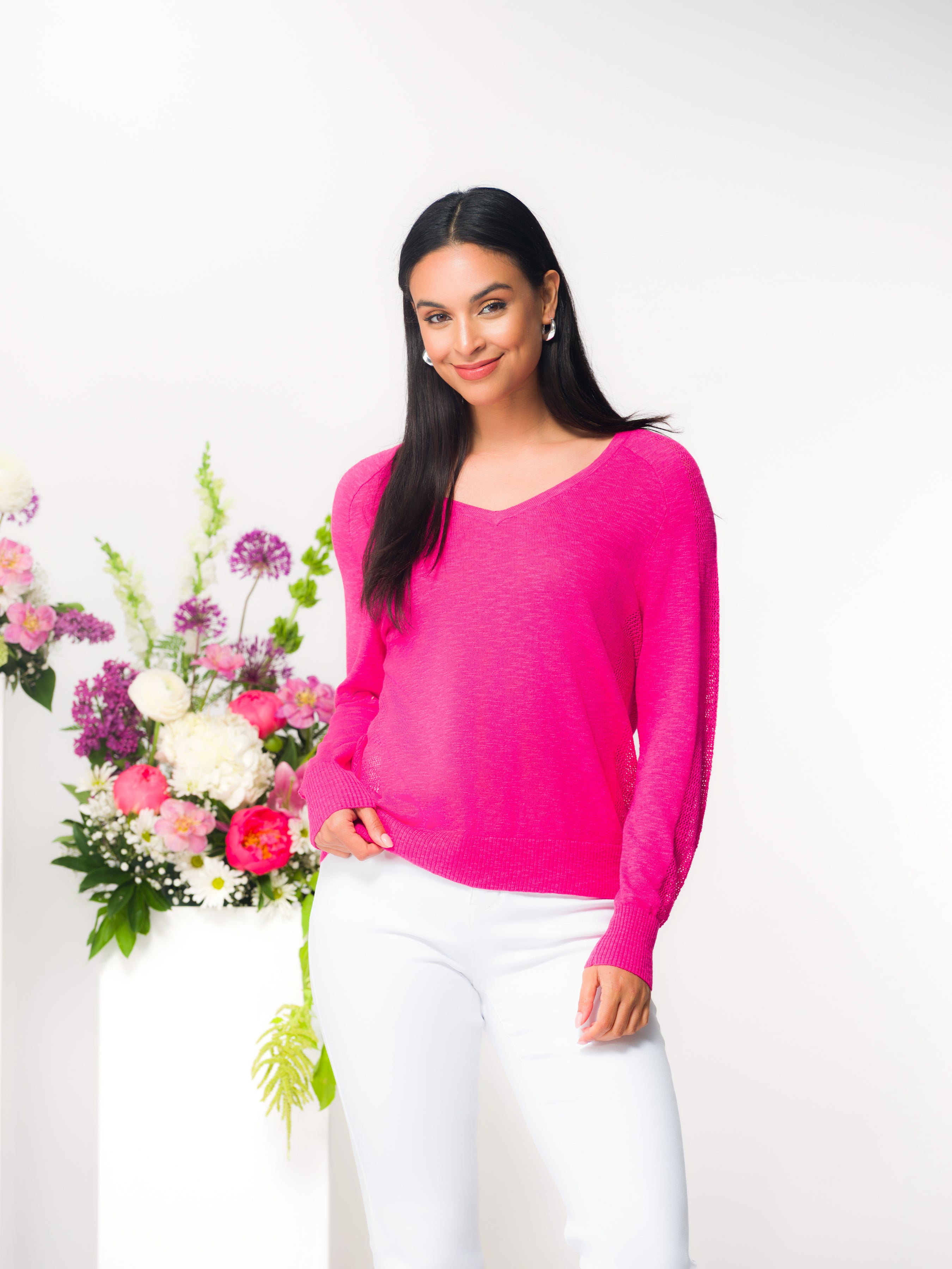 Woman wearing a pink lightweight V-neck knit sweater with long sleeves and a relaxed fit, paired with fitted pants. She stands in a studio setting beside a floral arrangement, with a soft, polished, and approachable overall look.