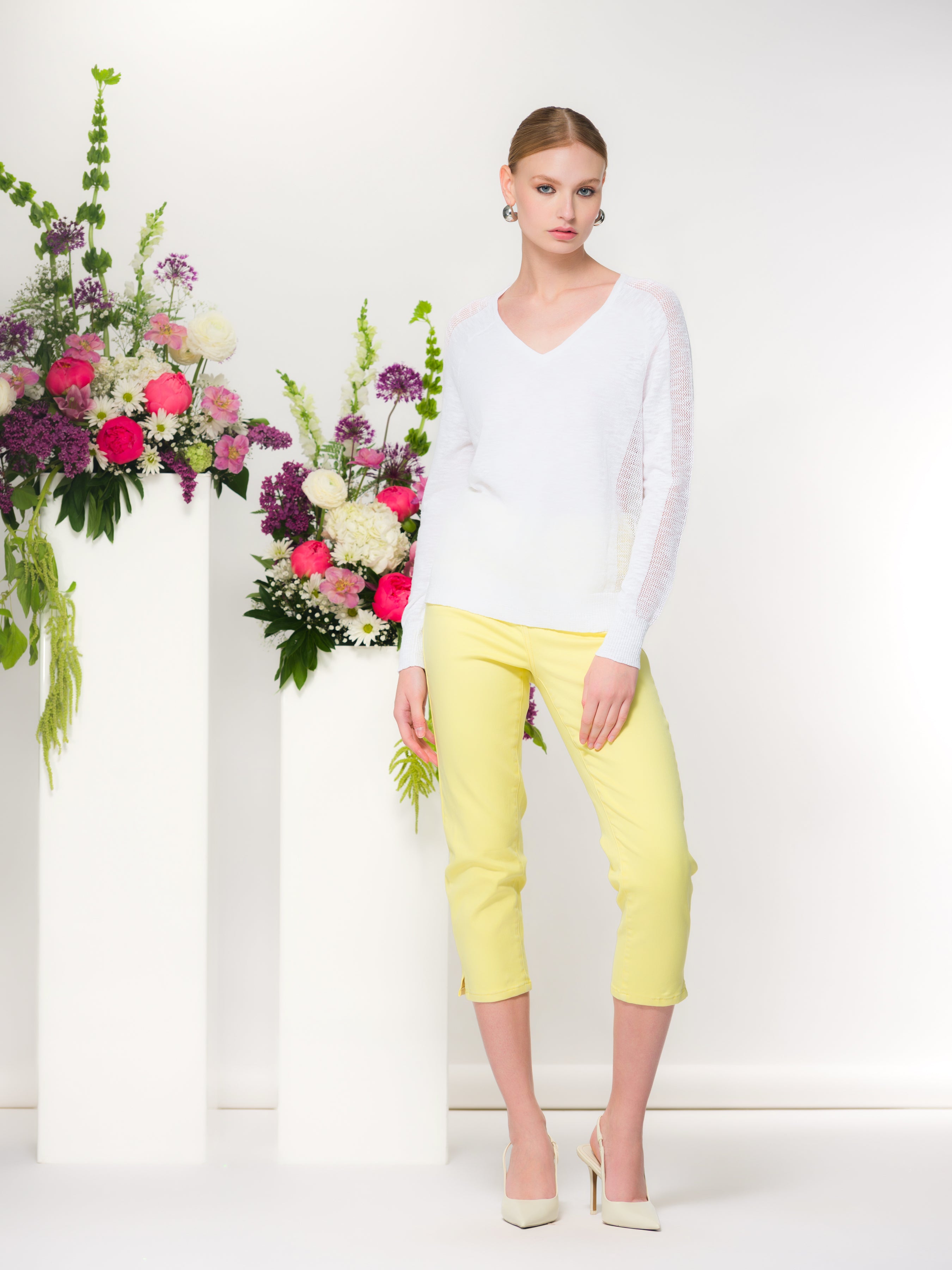 Woman wearing a white lightweight V-neck knit sweater with long sleeves and a relaxed fit, paired with fitted yellow capri pants. She stands in a studio setting beside a floral arrangement, with a soft, polished, and approachable overall look.