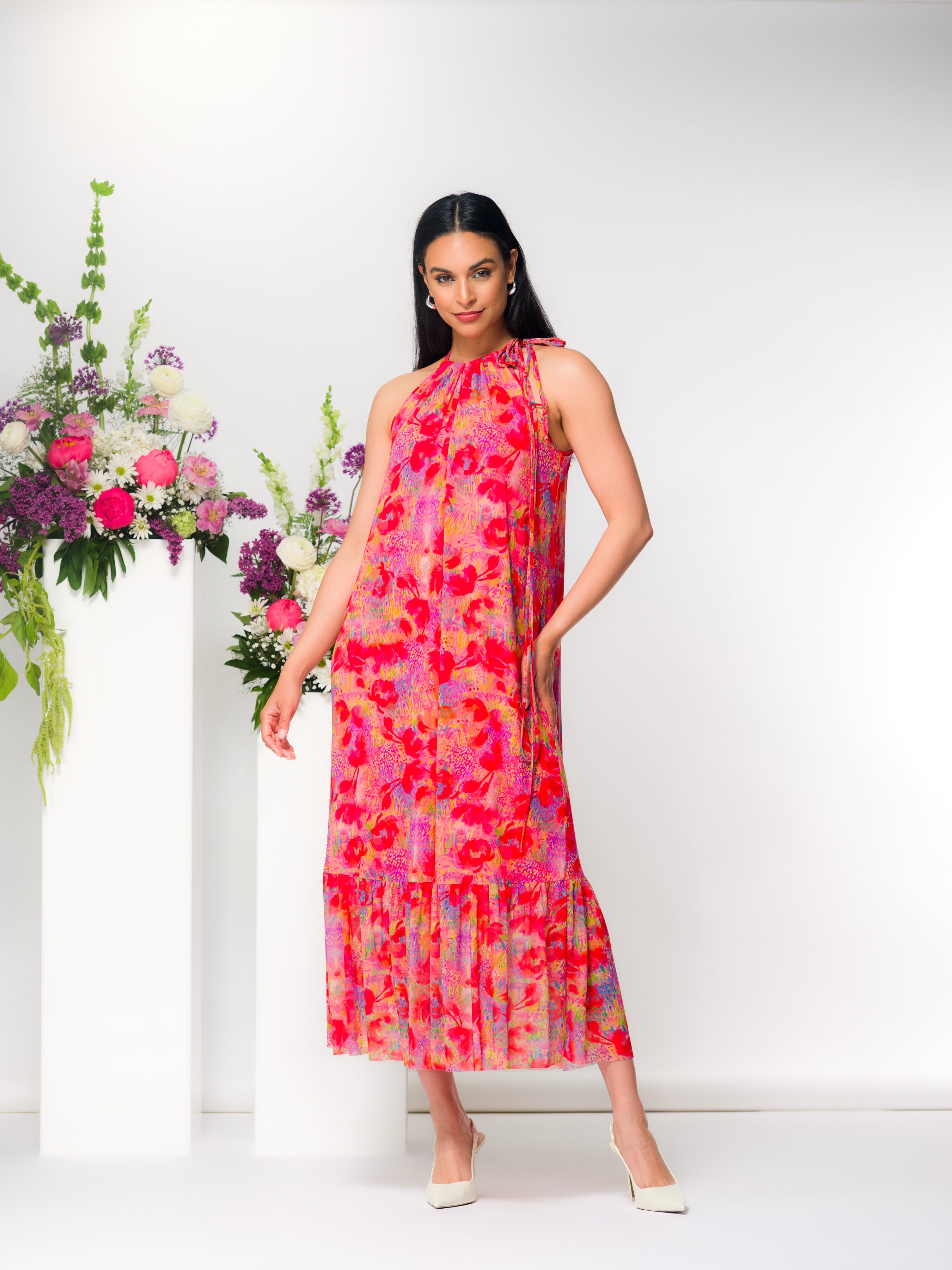 Model wearing a sleeveless halter-neck midi dress with a flowing, tiered skirt and soft drape, styled with heels.