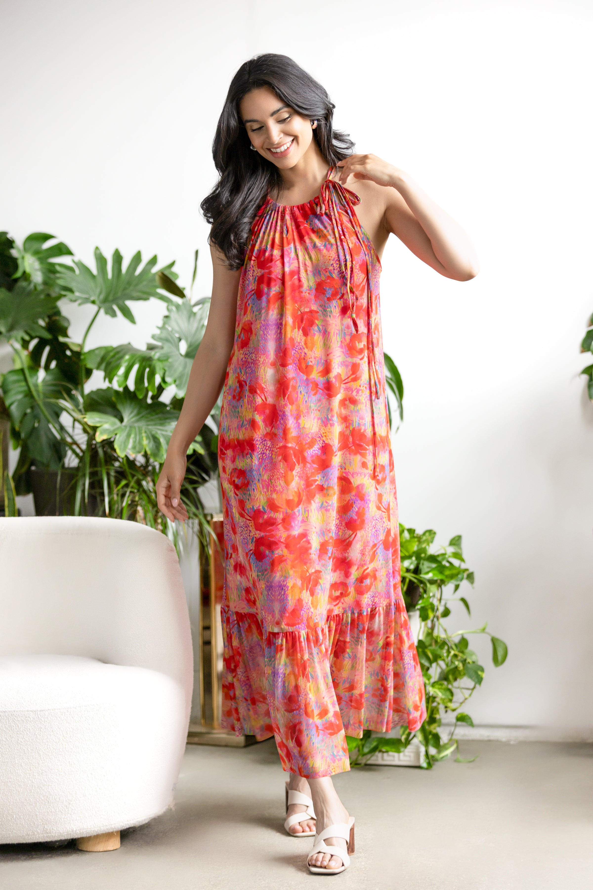 Model wearing a sleeveless halter-neck midi dress with a flowing, tiered skirt and soft drape, styled with heels.