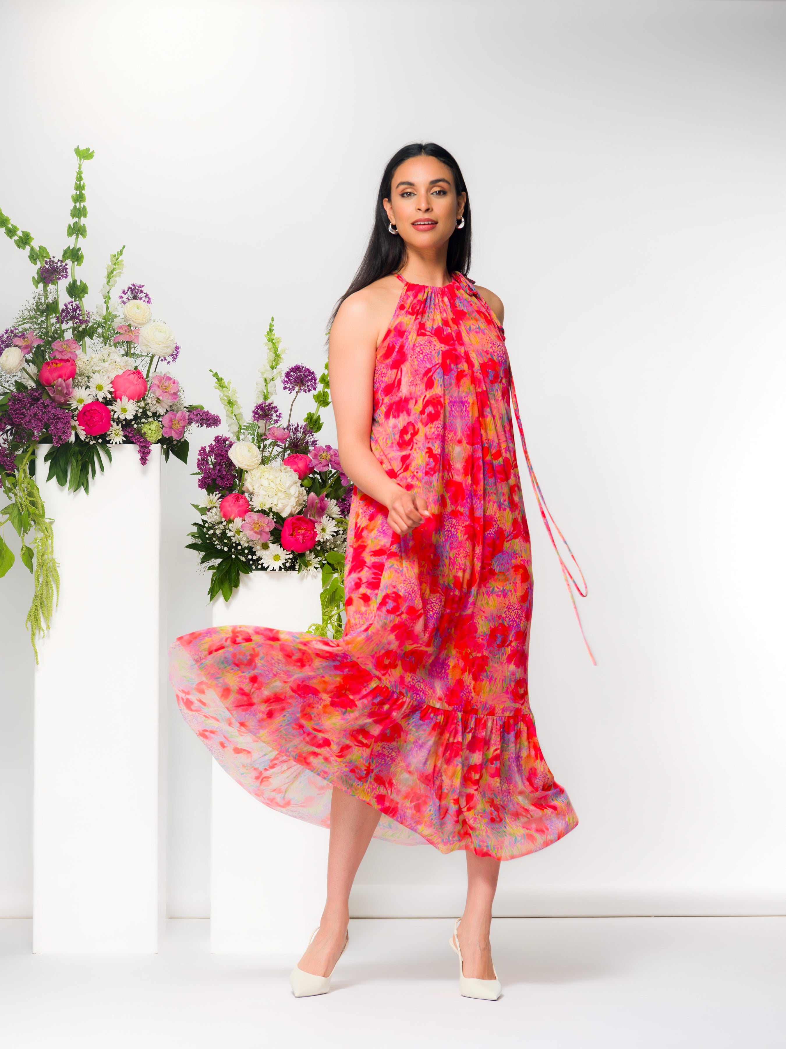 Model wearing a sleeveless halter-neck midi dress with a flowing, tiered skirt and soft drape, styled with heels.