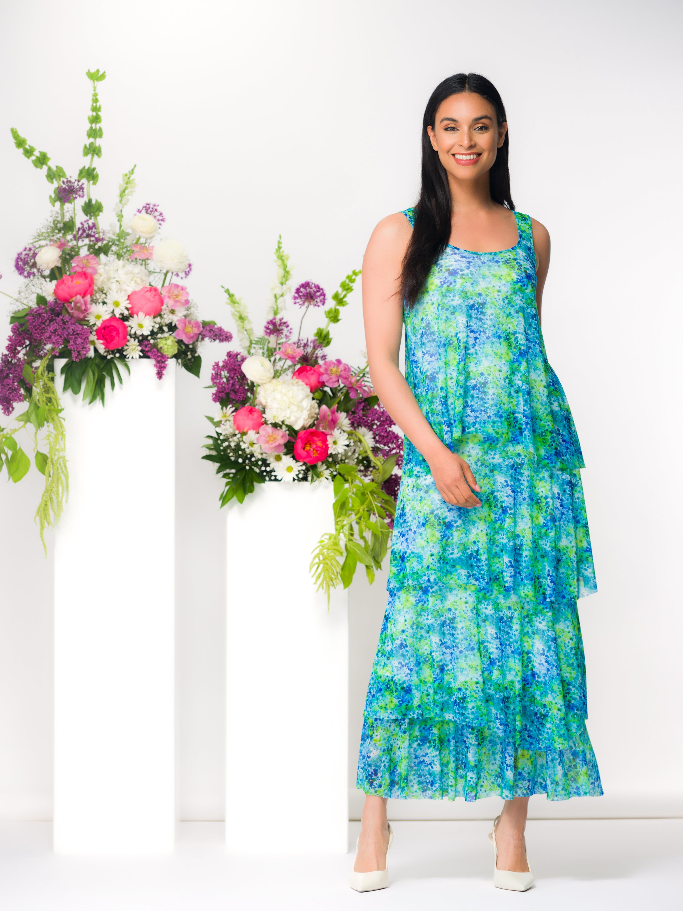 Sleeveless tiered midi dress with a flowing silhouette and floral pattern, shown on a model standing beside decorative floral arrangements.