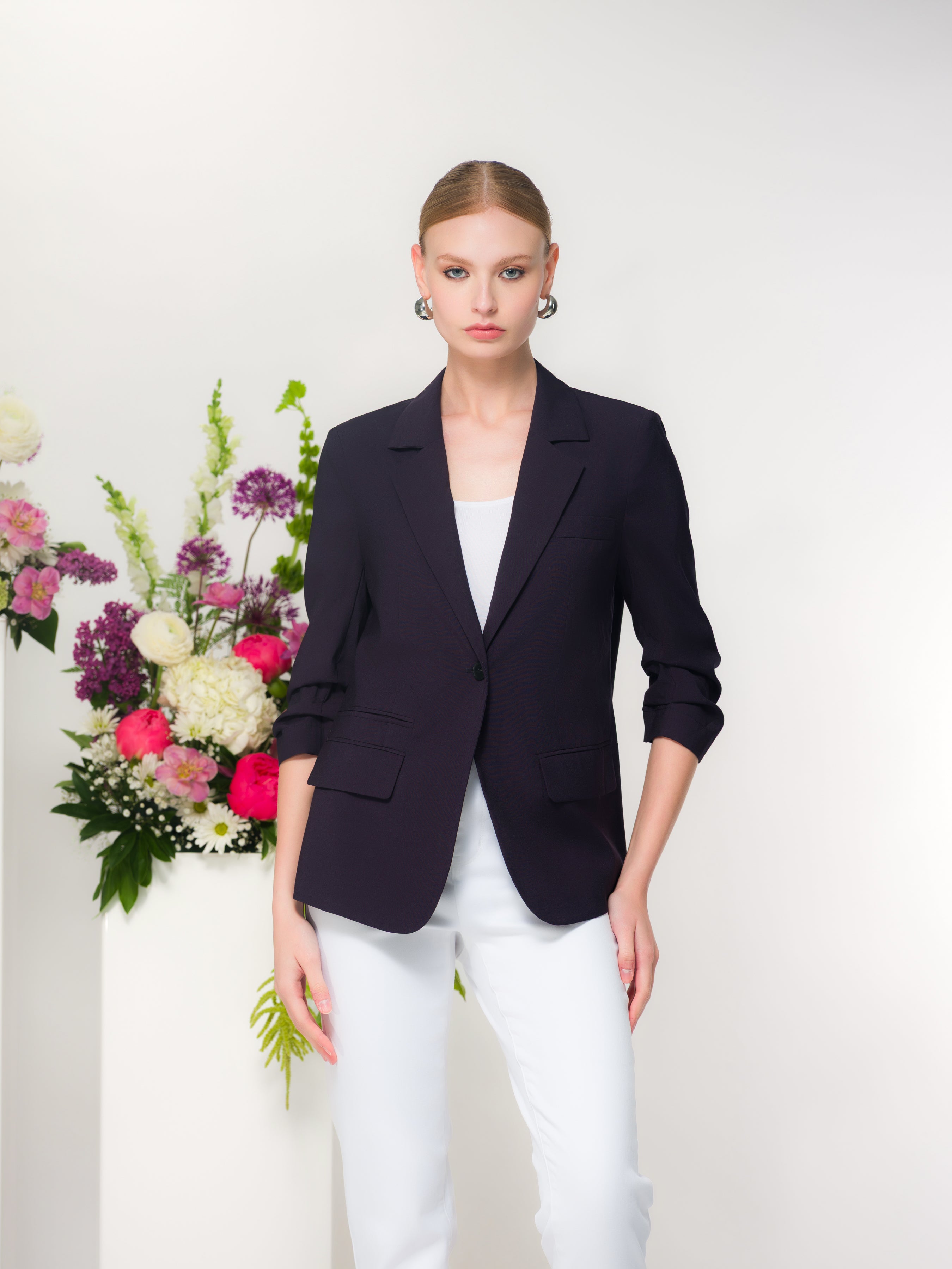 Women’s tailored blazer with a structured silhouette and subtle stretch, styled with a simple top and slim-fit pants, photographed on a model standing against a minimal studio backdrop with floral accents.