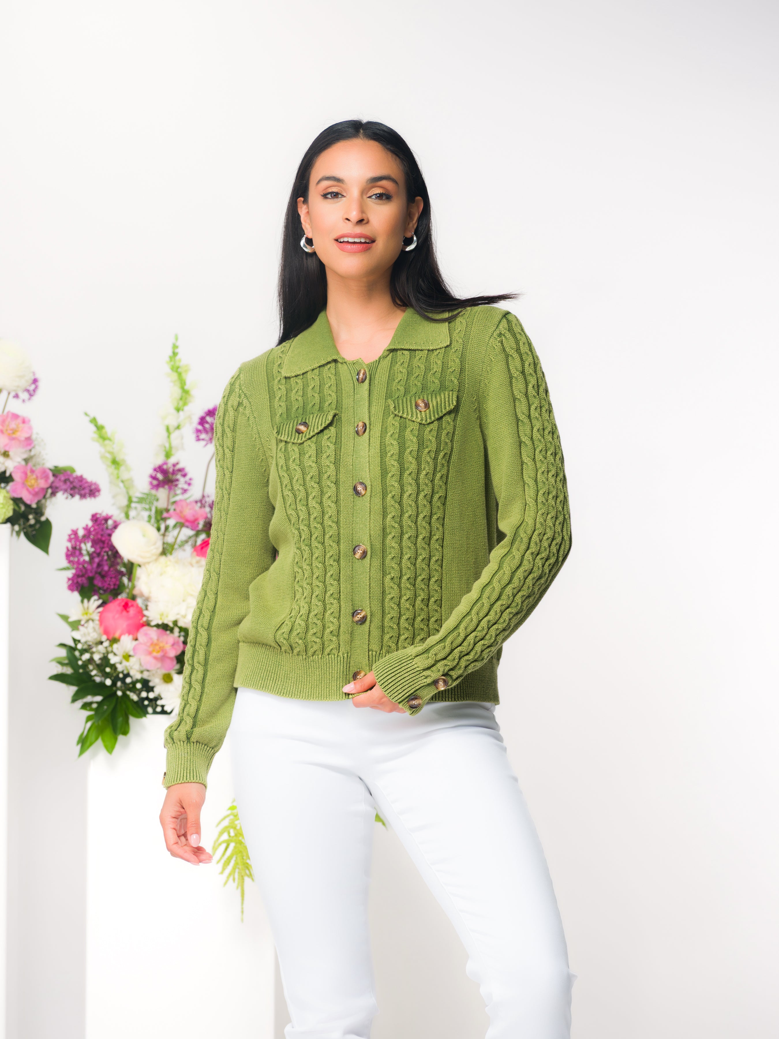 Model wearing a women’s cable knit button-front cardigan with a collared neckline, long sleeves, and textured detailing, paired with fitted pants, photographed in a studio setting with floral accents.